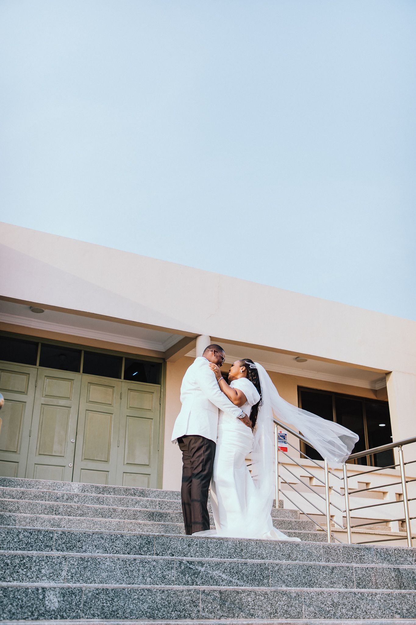 Bride and groom in white wedding attire share a romantic kiss on grand stone steps outdoors.