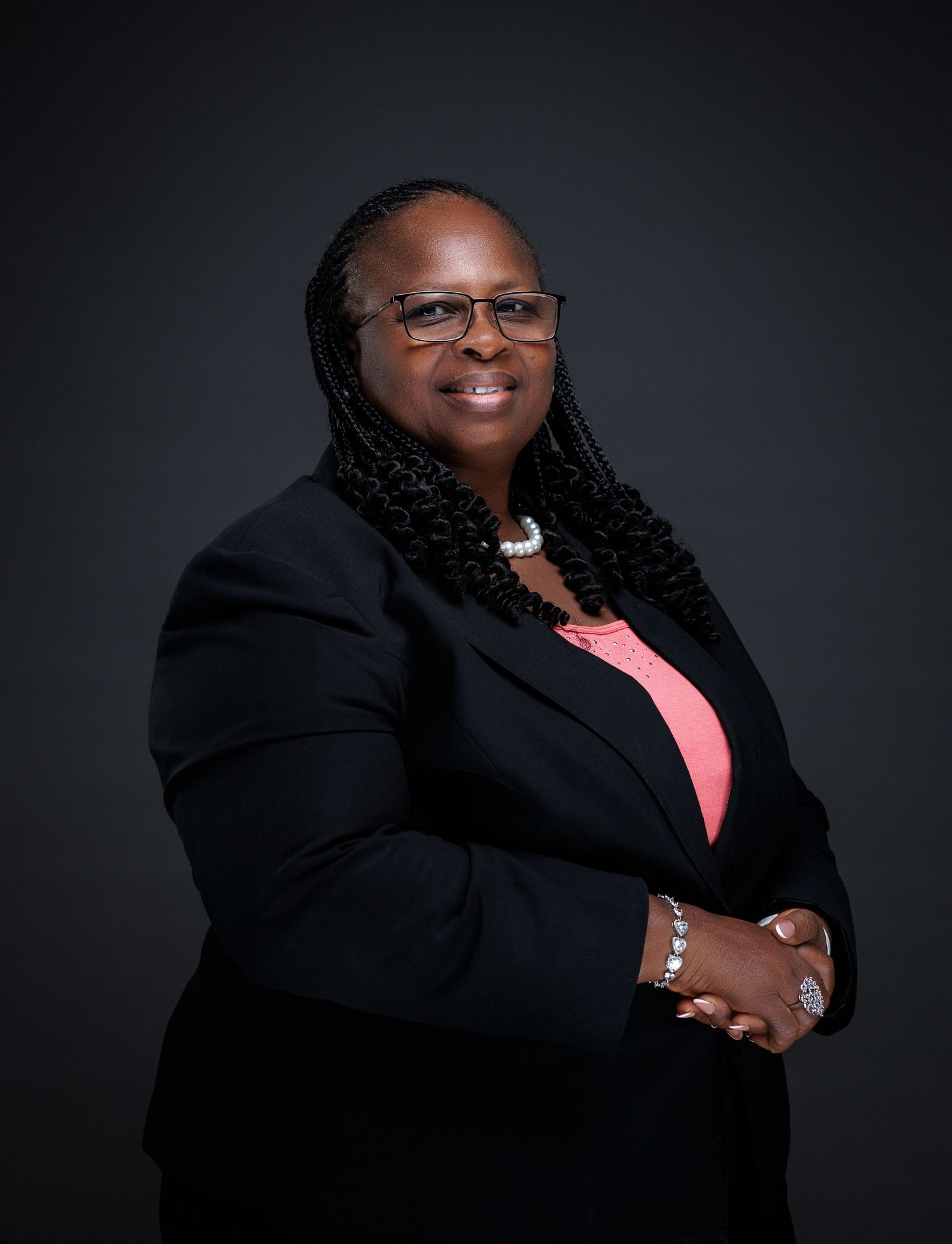 Professional woman in black blazer with braids and glasses smiling in corporate studio portrait.