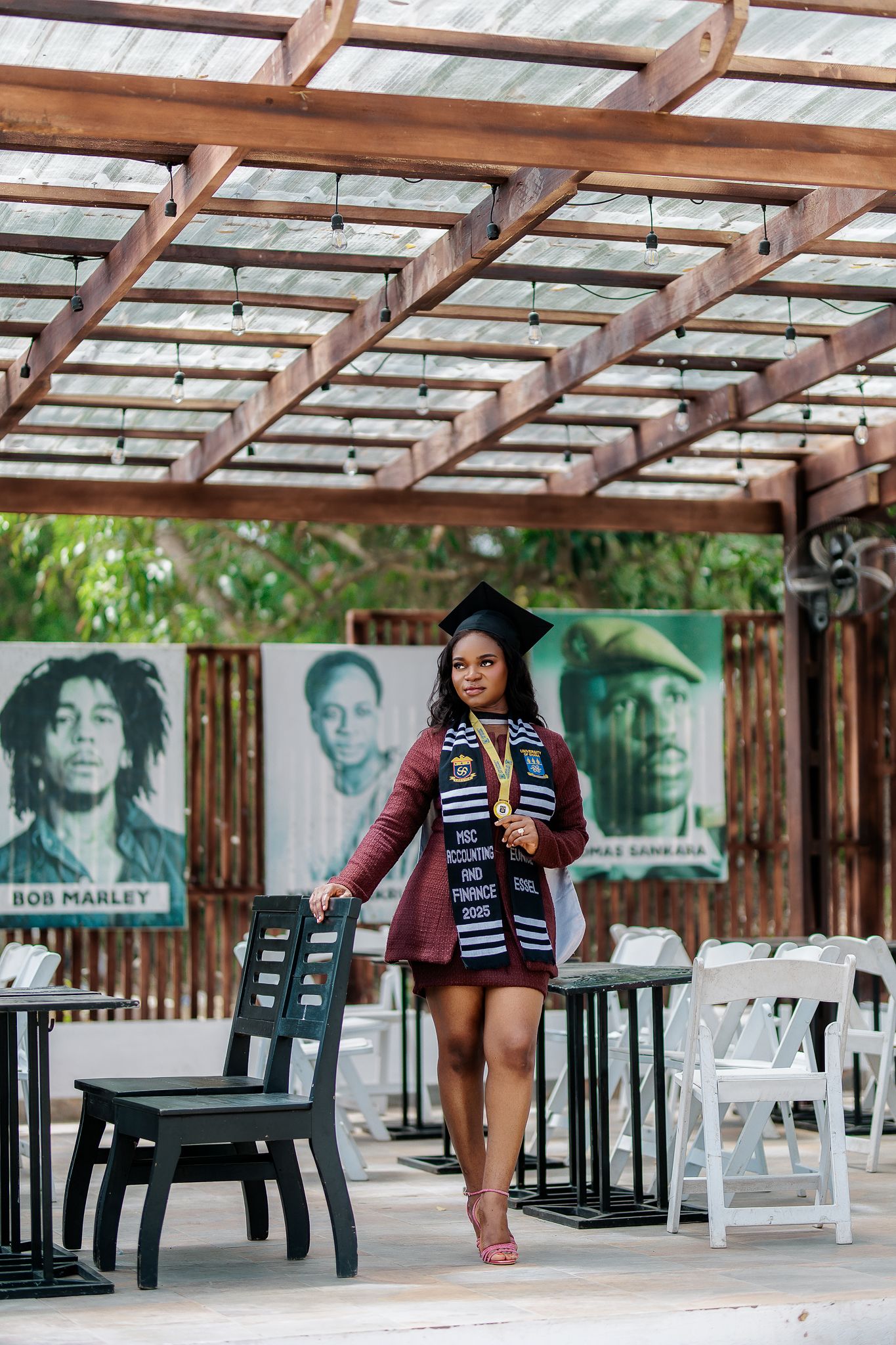 Black female graduate in cap and gown with stole, posing under pergola near Bob Marley mural.