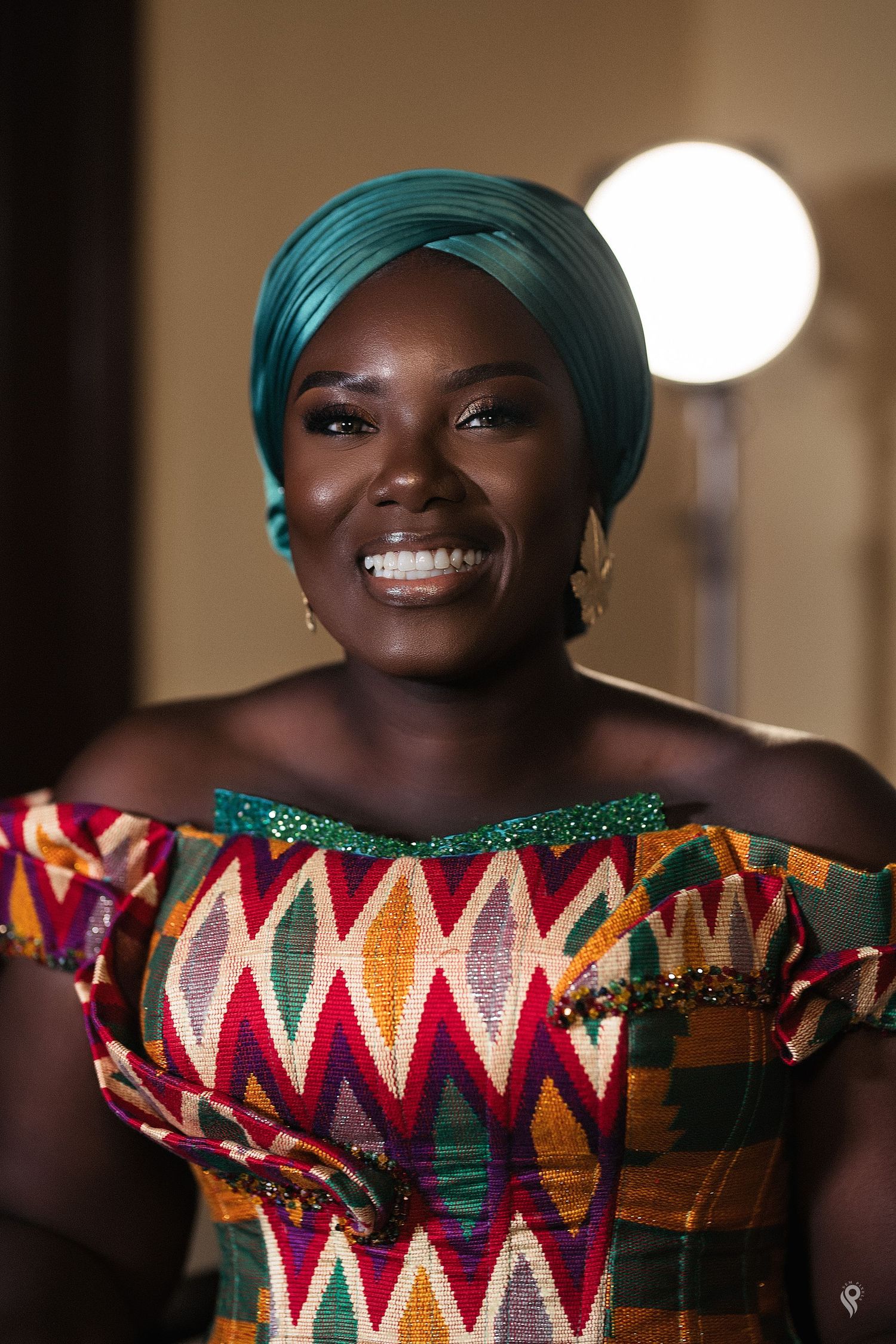 Smiling African woman in vibrant kente off-shoulder dress and teal head wrap under studio lighting.