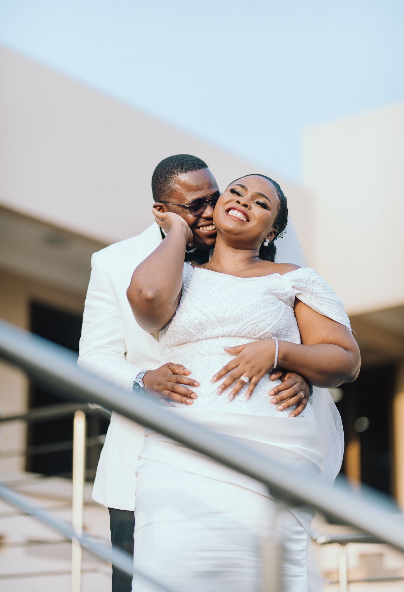 Groom in white suit embraces smiling bride in white off-shoulder gown on sunlit outdoor steps.