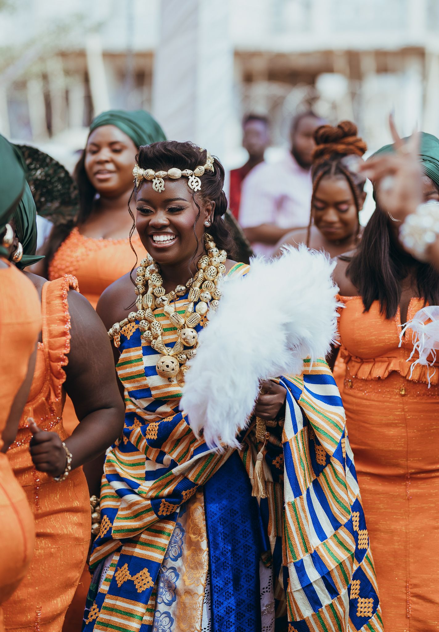 Bride in colorful Kente cloth and gold jewelry smiling at traditional Ghanaian wedding ceremony.