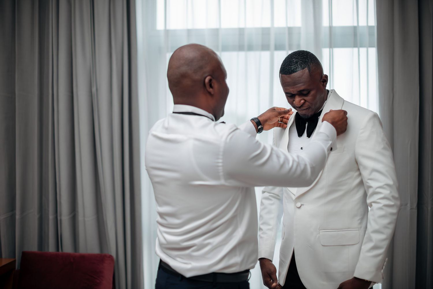 A groomsman helps a groom in a white tuxedo adjust his bow tie in a hotel room.