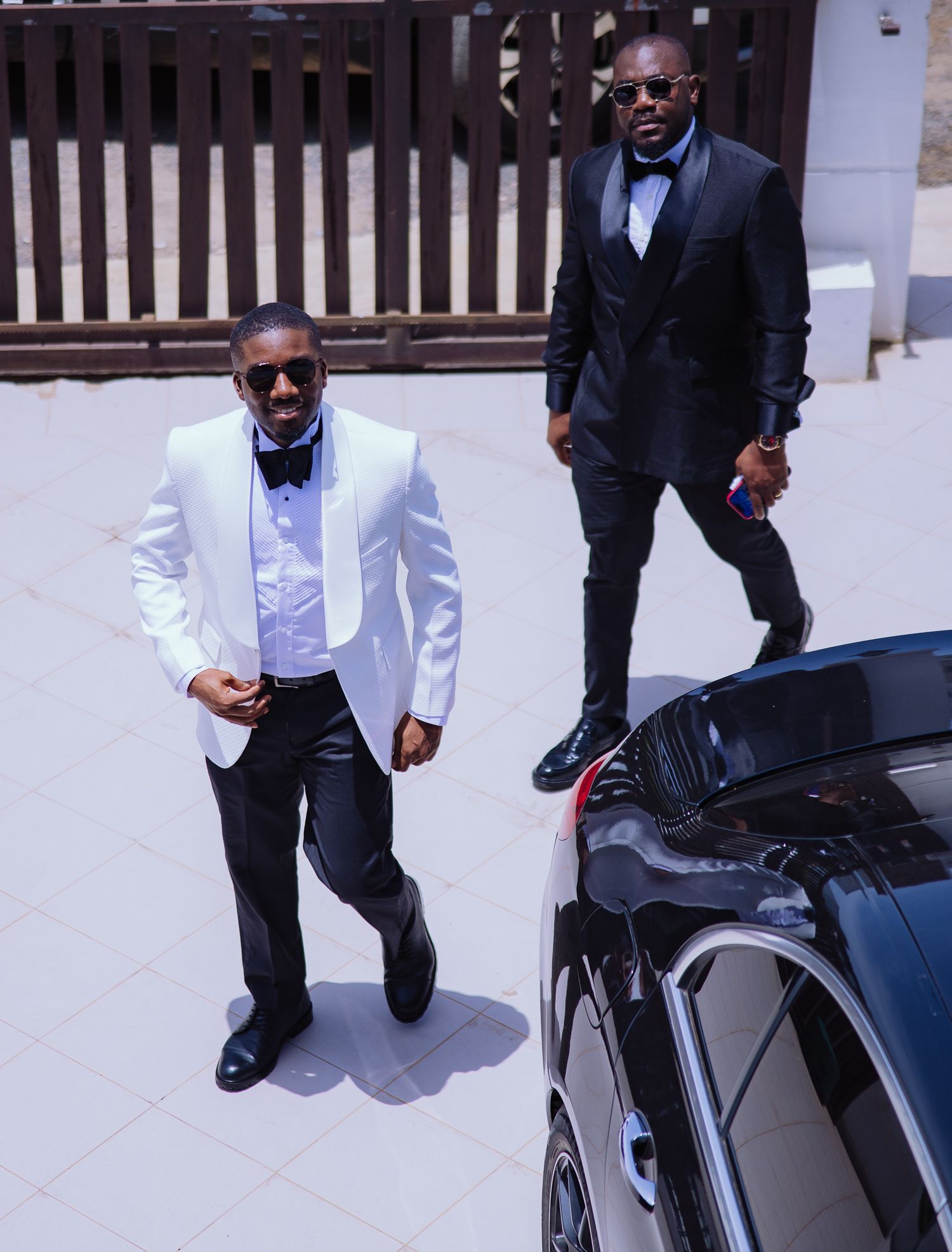 Two stylish groomsmen in black and white tuxedos walking confidently beside luxury black car.