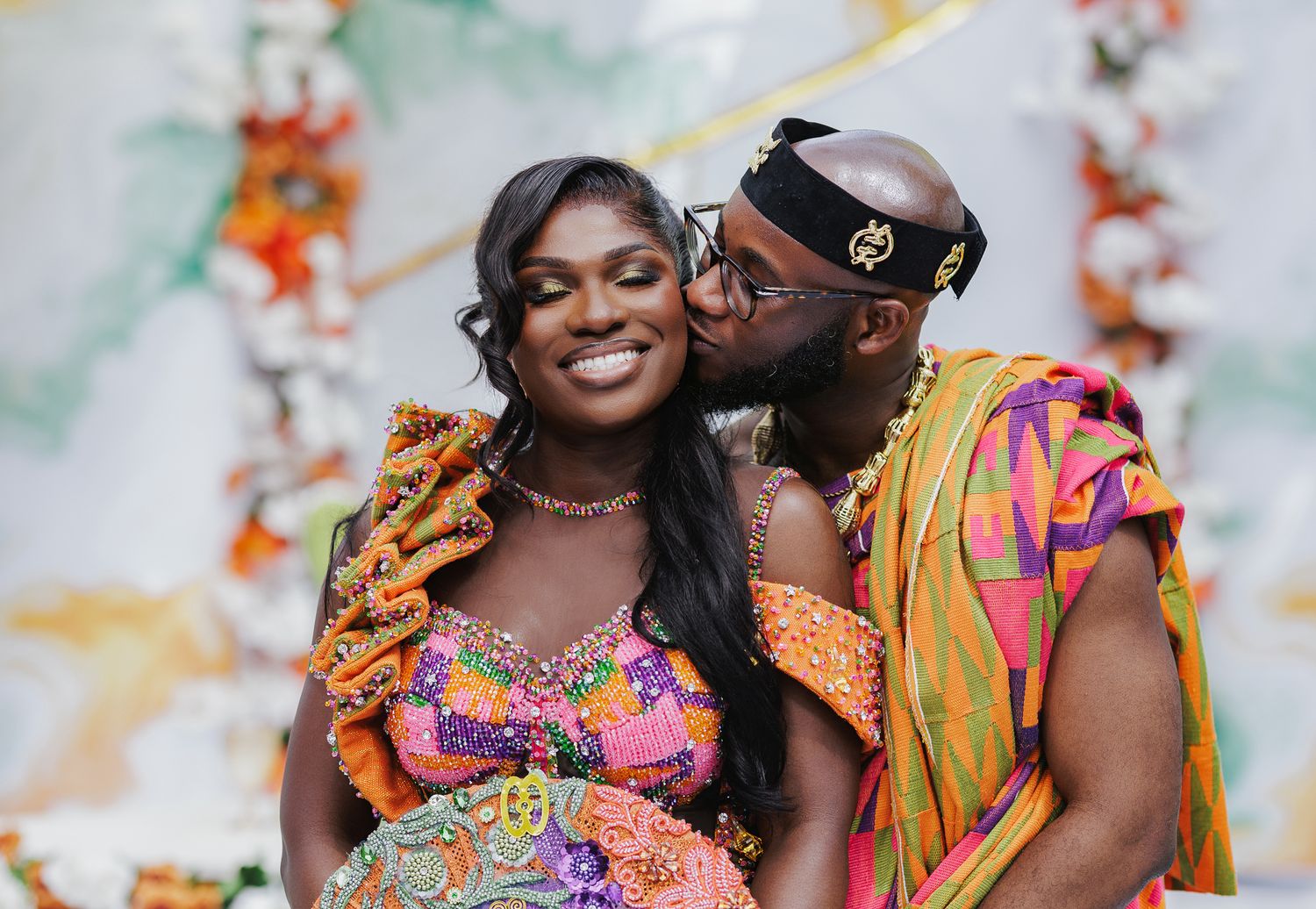 A groom in colorful Kente attire kisses his bride on the cheek during a traditional African wedding.