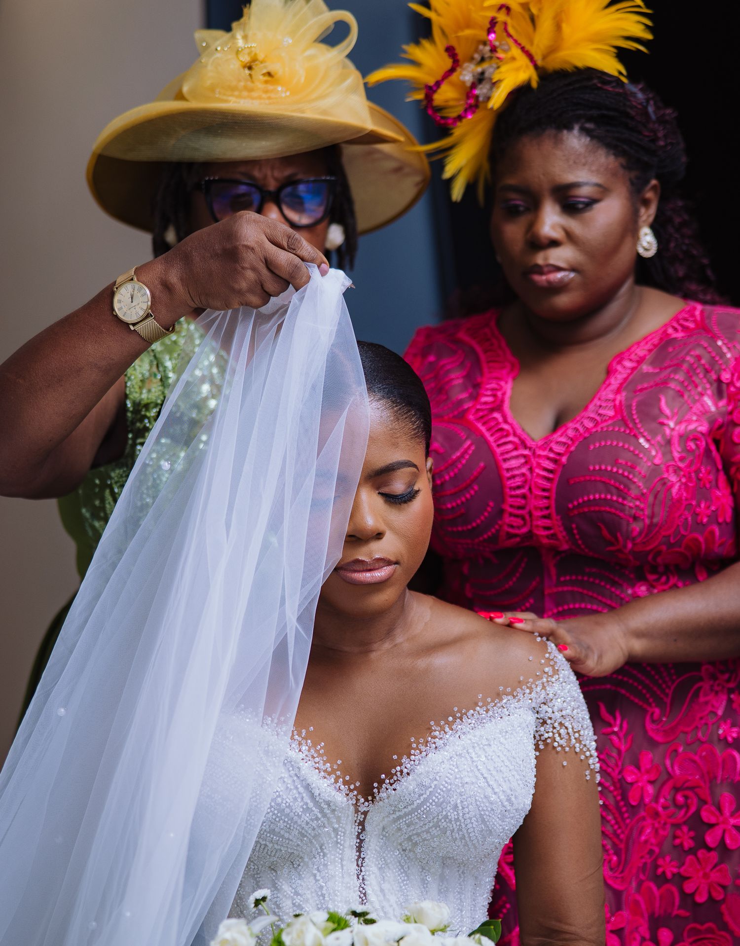 Mother adjusts daughters wedding veil while attendant in pink dress looks on.