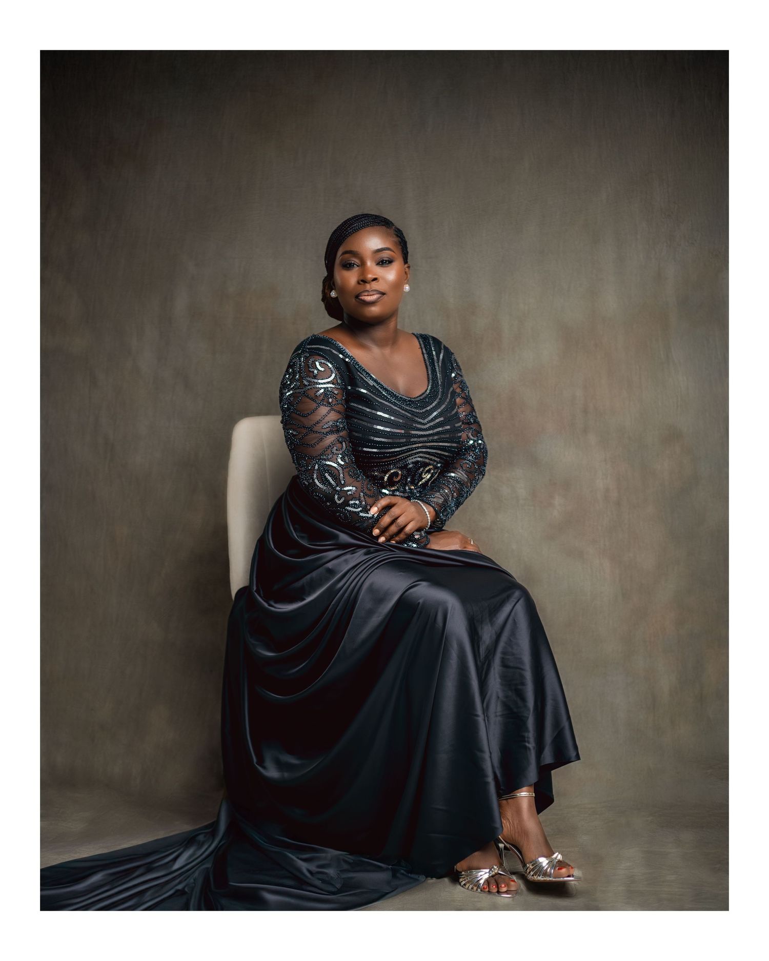 Elegant Black woman in navy beaded long-sleeve gown seated on white chair in studio setting.