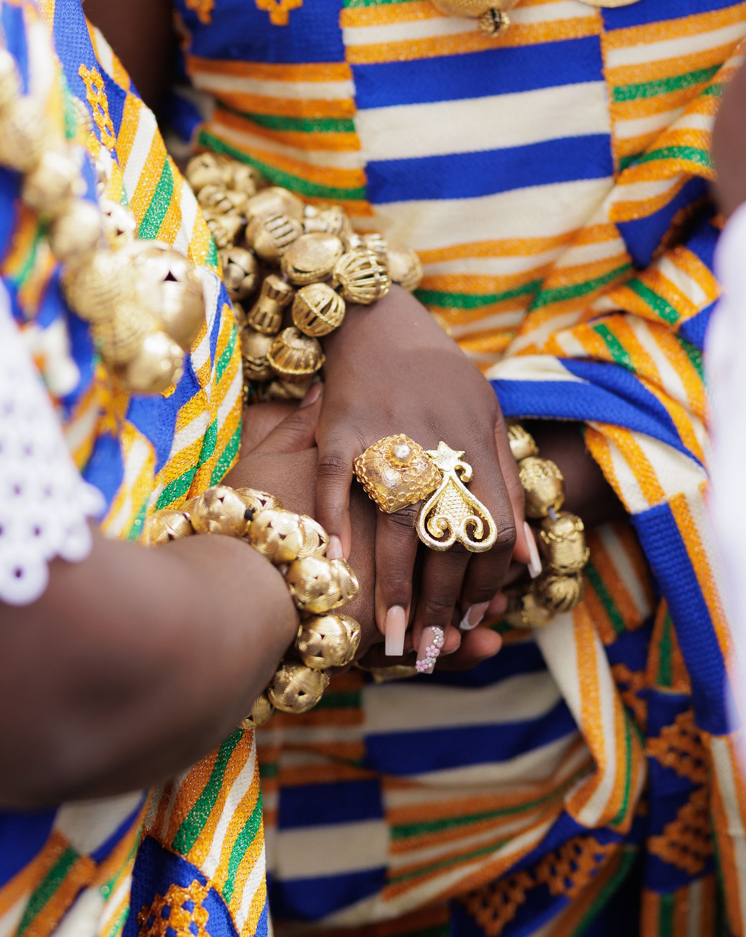Close-up of hands adorned with gold beaded bracelets and a decorative ring over Kente fabric.