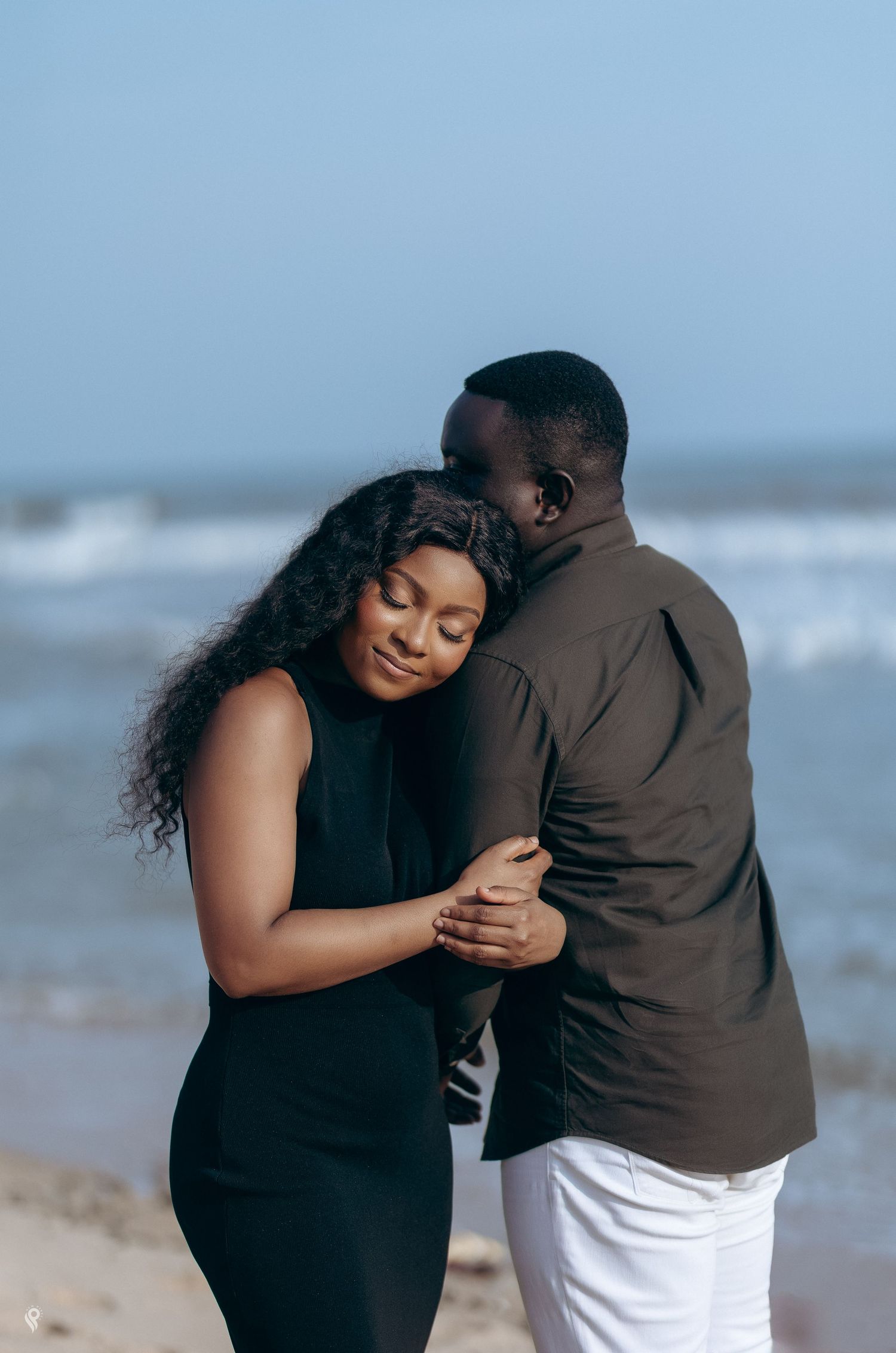 Tender Black couple embracing on a beach, woman in black dress leaning into mans shoulder.