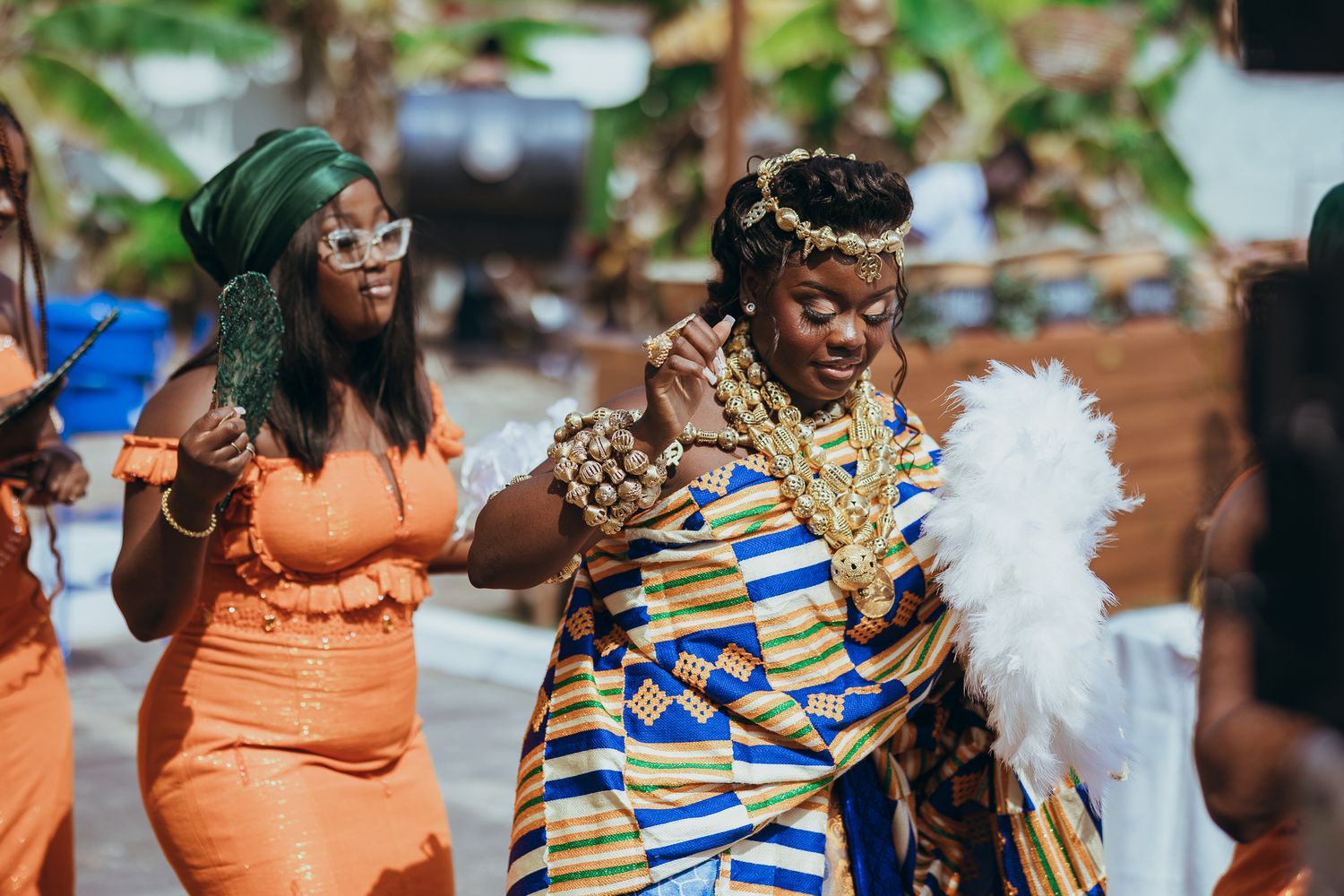 Bride in Kente cloth dancing with guest in orange outfit at Ghanaian traditional wedding.