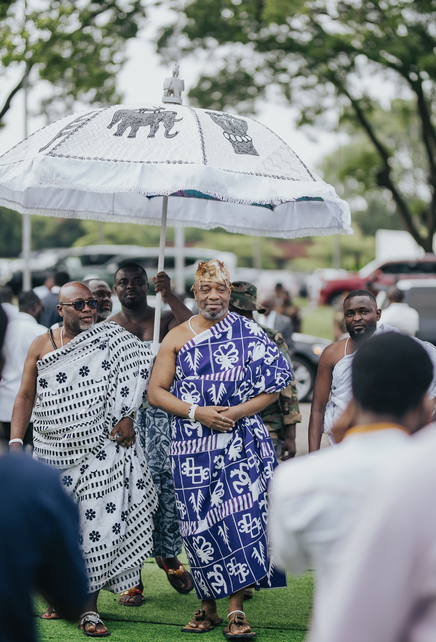 Ghanaian chief in blue kente cloth standing under white ceremonial elephant umbrella.