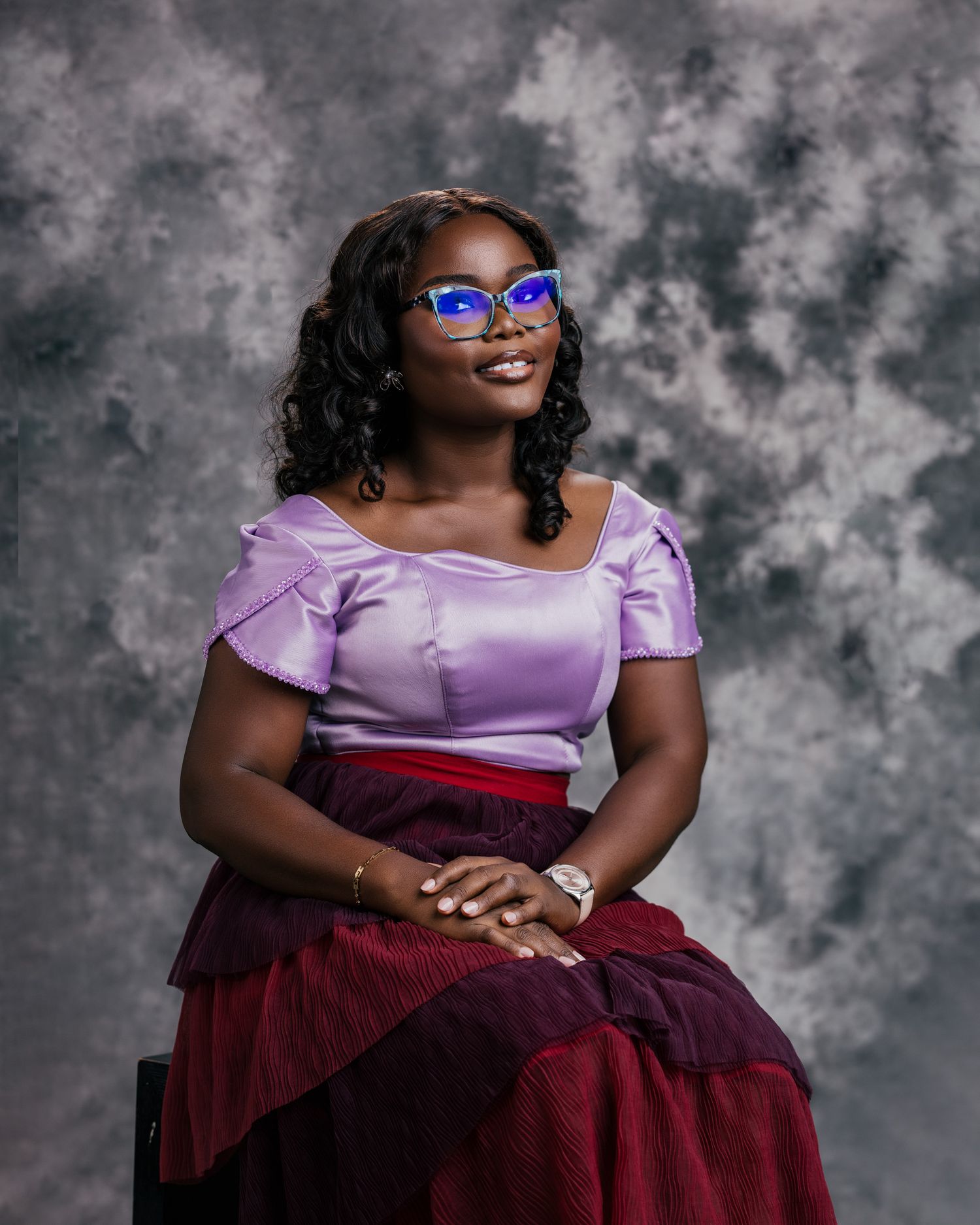 Young woman in purple satin off-shoulder top and burgundy skirt wearing blue cat-eye glasses, seated in studio.