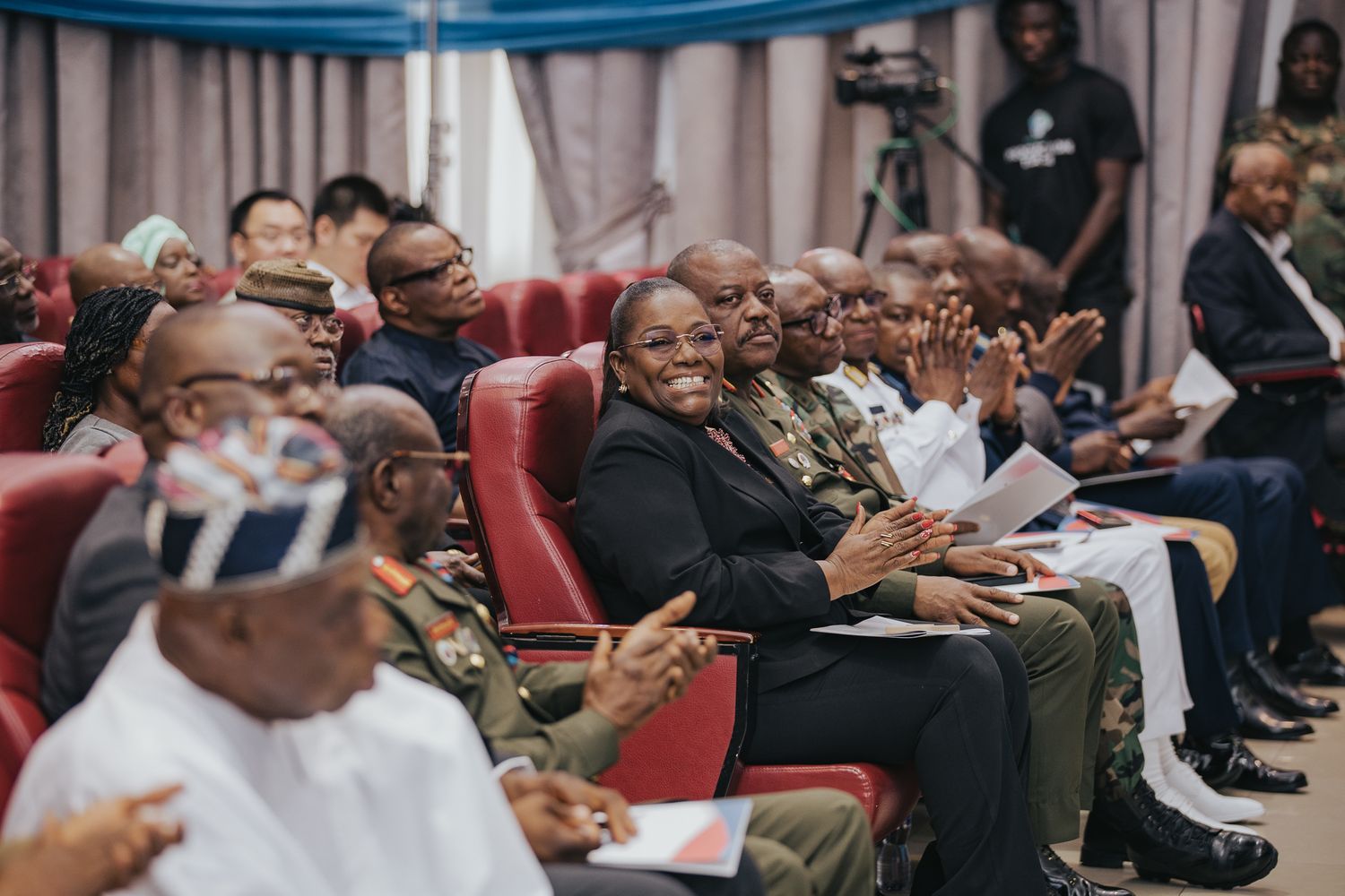 Group of military and civilian attendees smiling and applauding at a formal indoor conference.