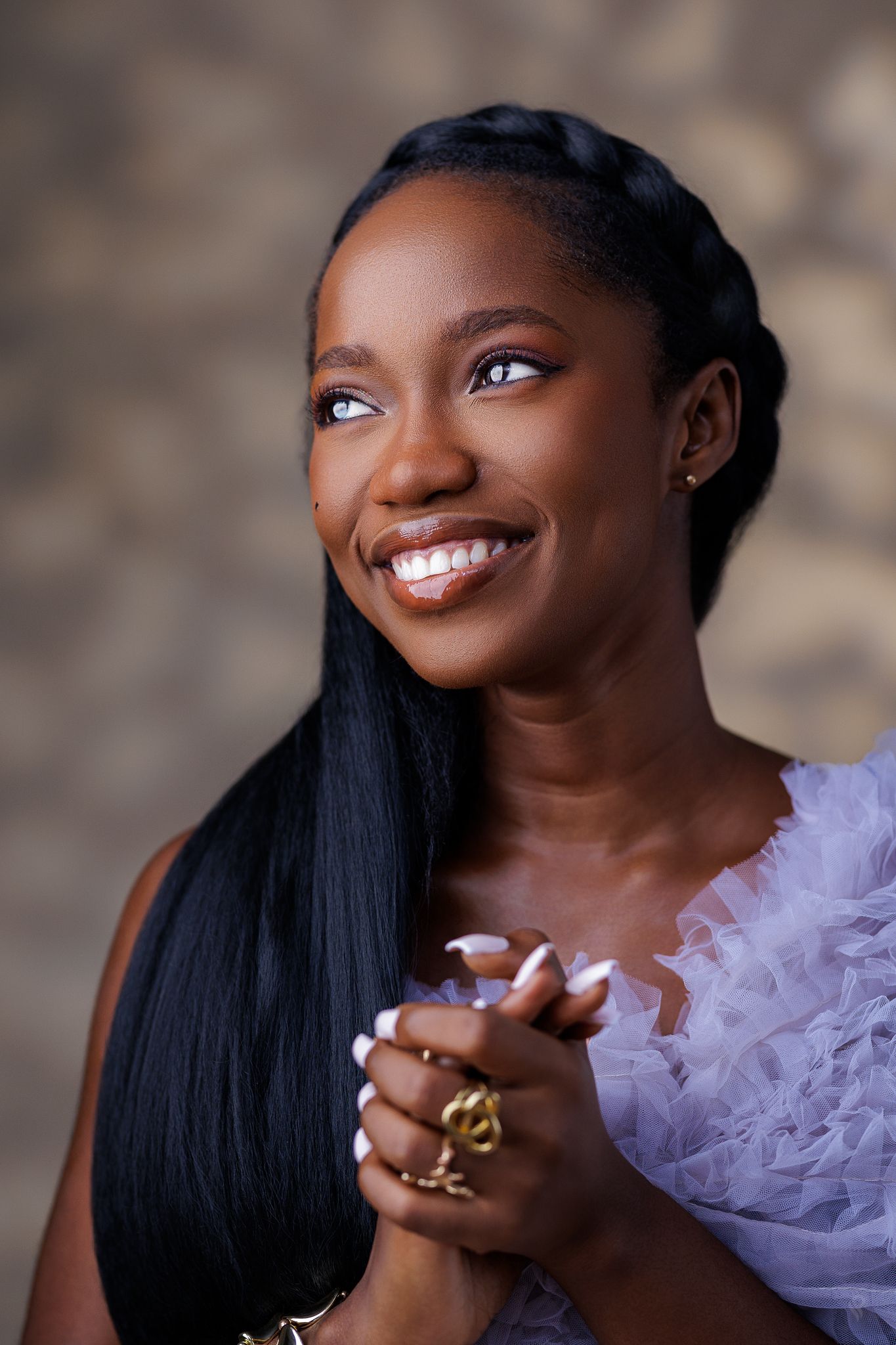 Smiling Black woman in lavender ruffled dress with braided hair and gold rings, clasping hands.