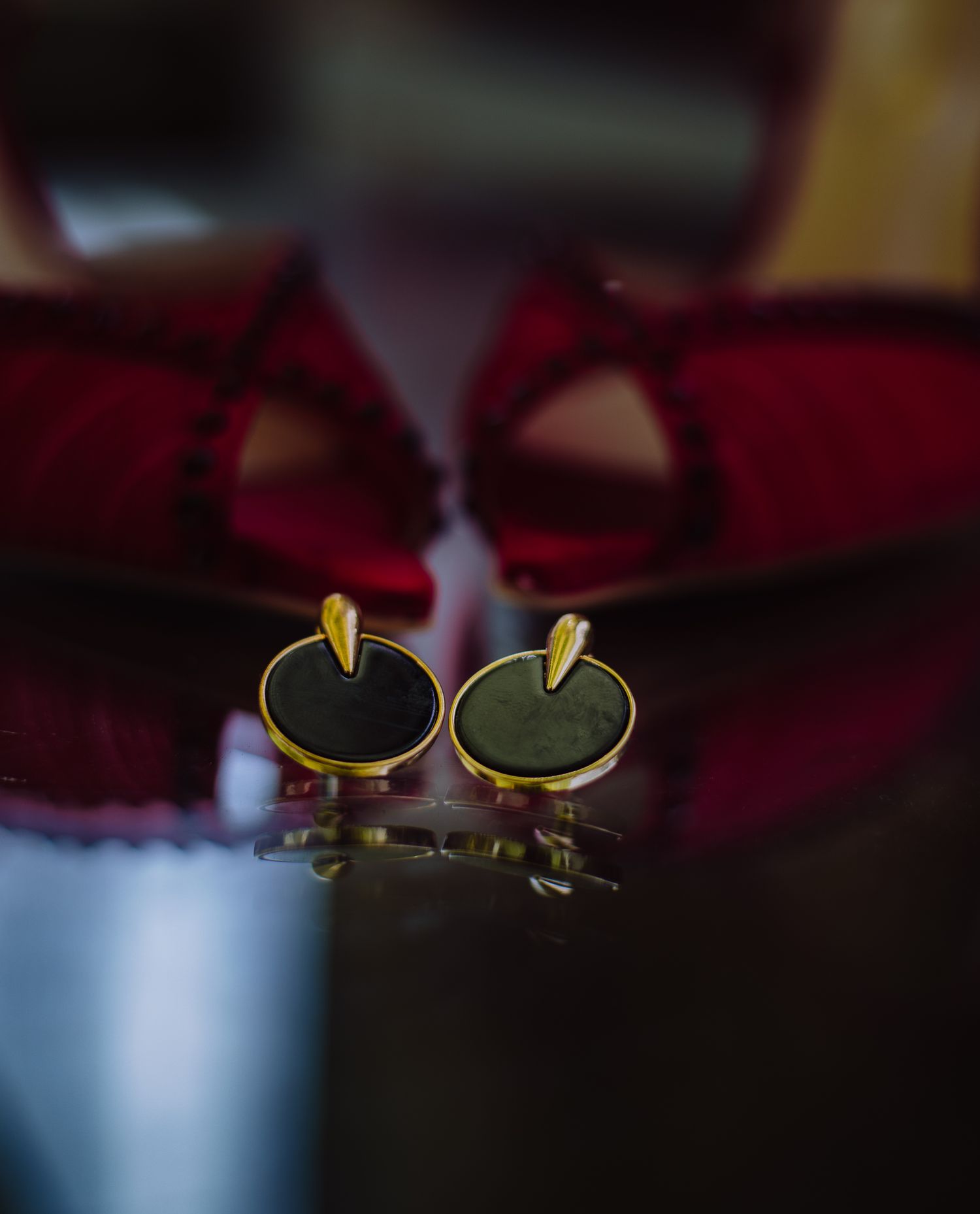 Elegant black and gold circular cufflinks displayed before red velvet wedding shoes on reflective surface.