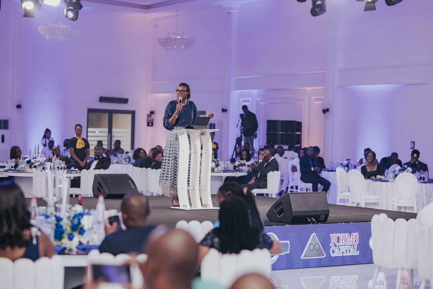 Confident woman in white outfit addresses large audience at a formal corporate banquet event.