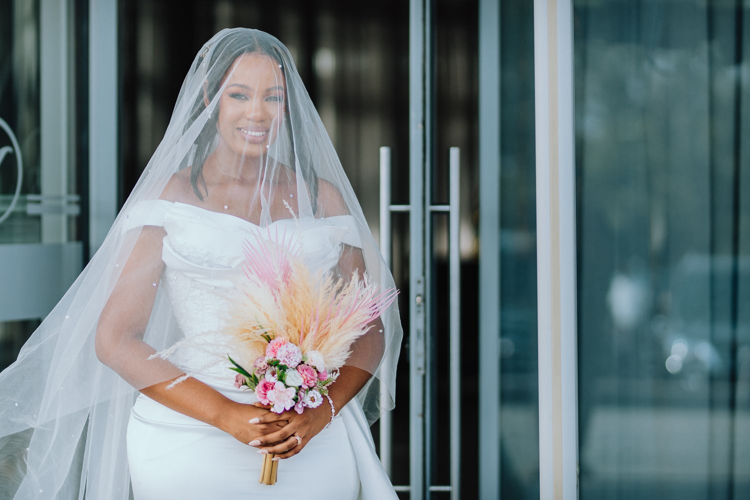 A smiling Black bride in a white off-shoulder gown holds a floral bouquet while wearing a long veil.