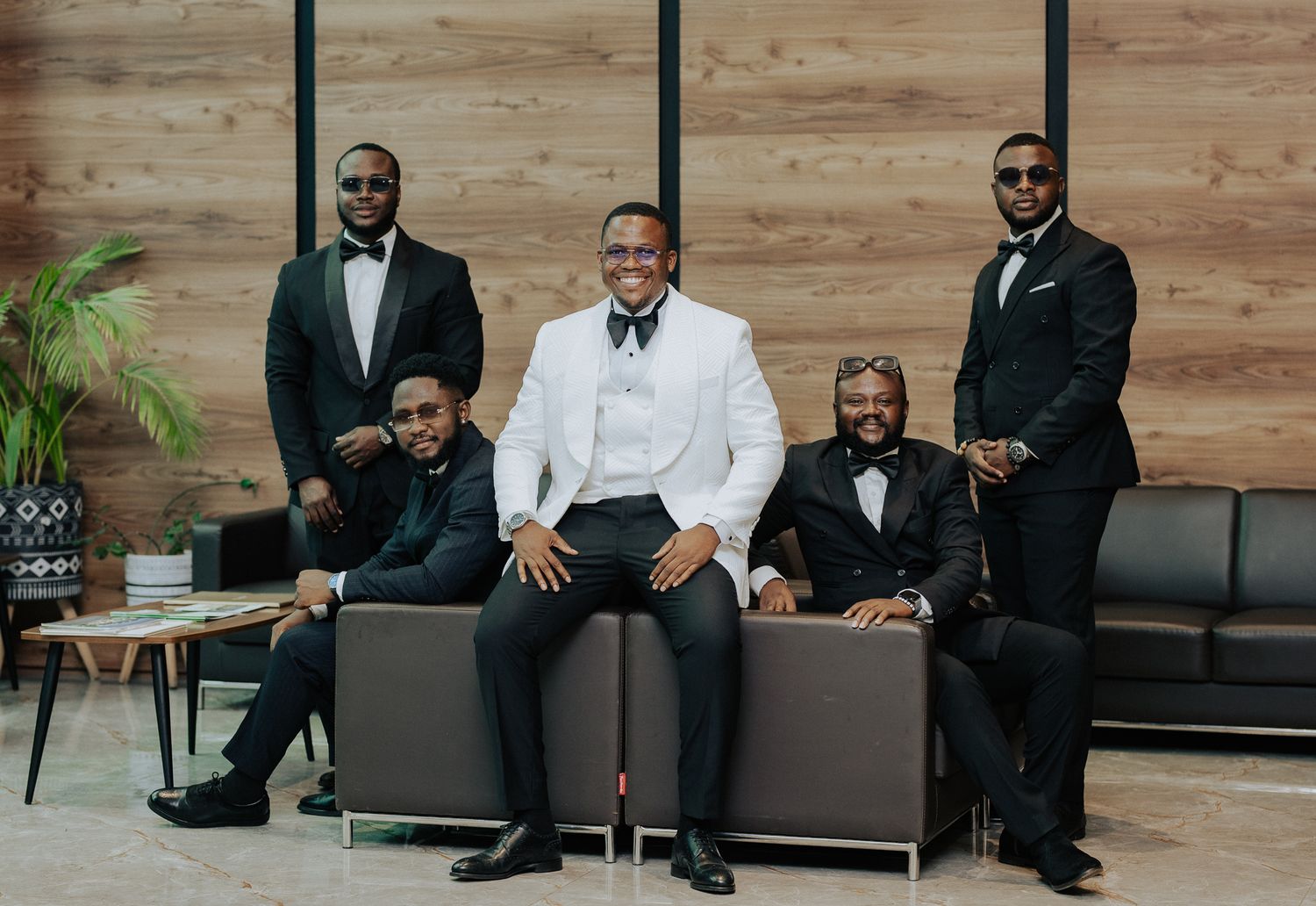 A groom in white tuxedo poses with four groomsmen in black suits in a modern lobby.