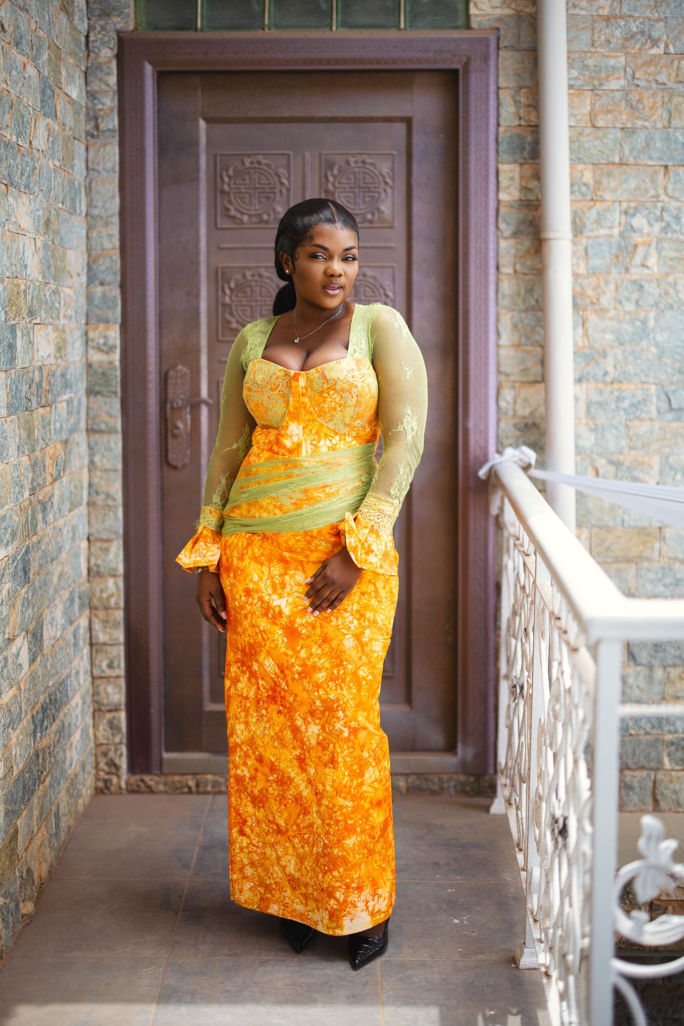 A woman in a vibrant orange and green traditional African lace gown stands by a doorway.