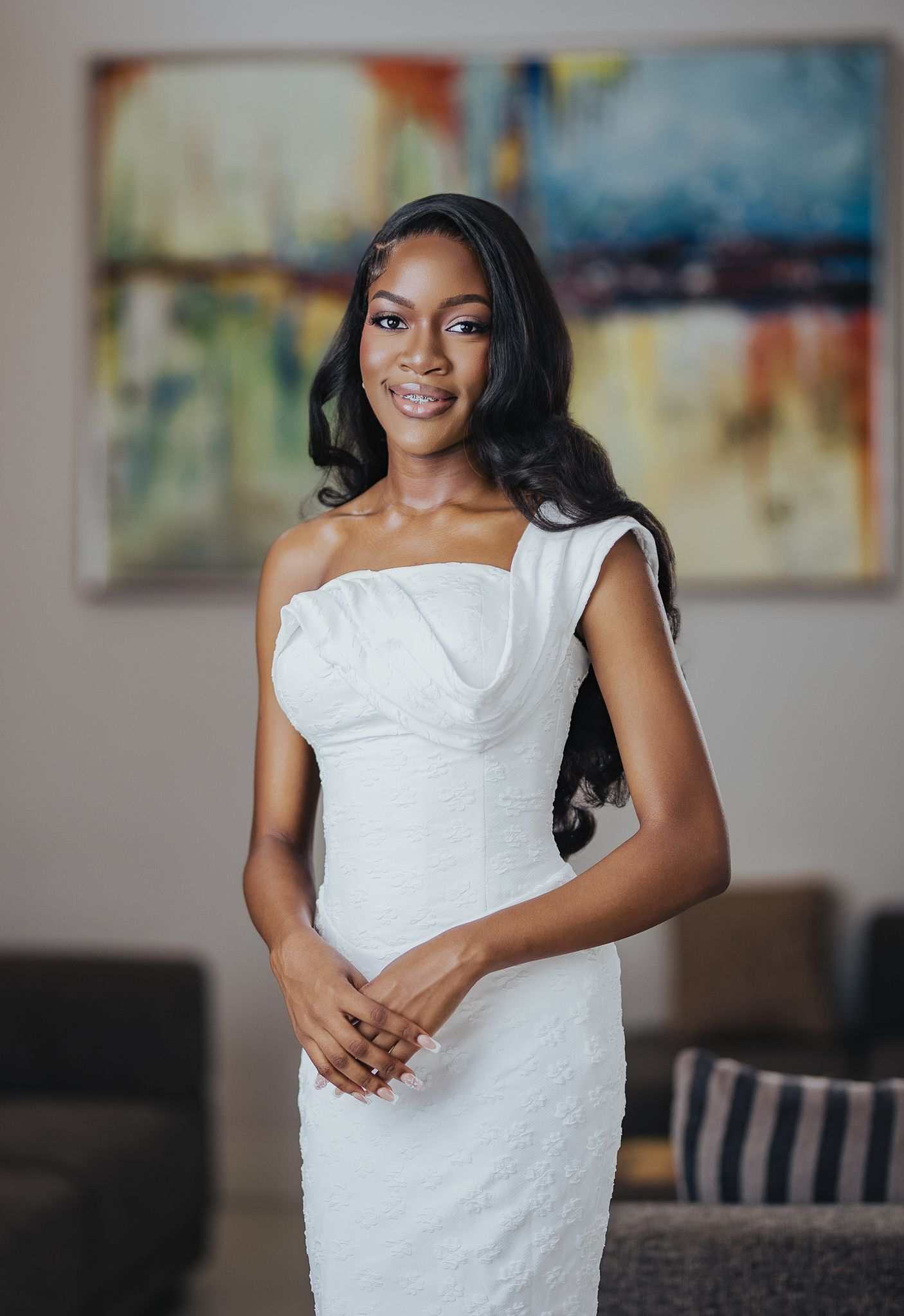 Tall elegant woman in white one-shoulder fitted dress standing indoors with colorful artwork in background.