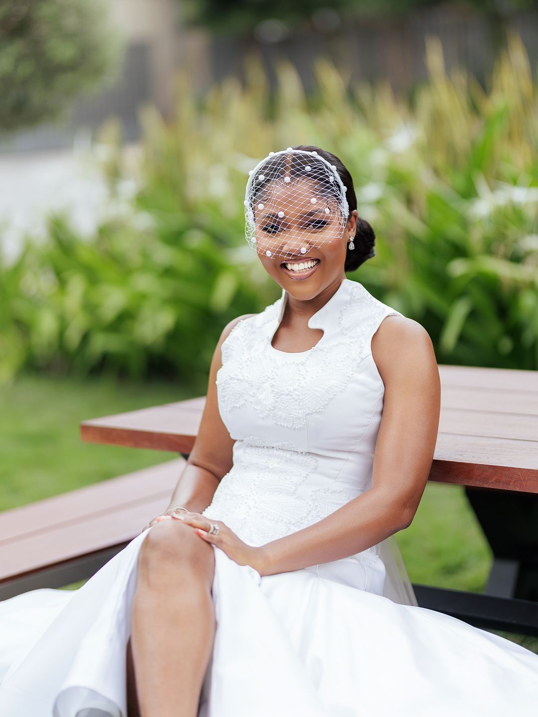 Smiling bride in white lace wedding dress and birdcage veil sitting outdoors at a wooden bench.