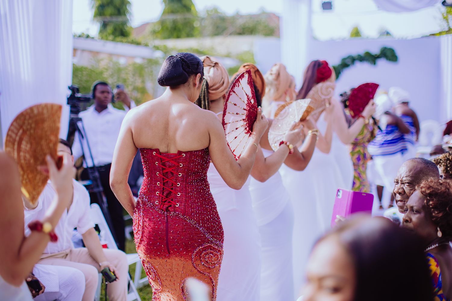 Women in vibrant traditional African attire dance joyfully with decorative fans at an outdoor wedding celebration.