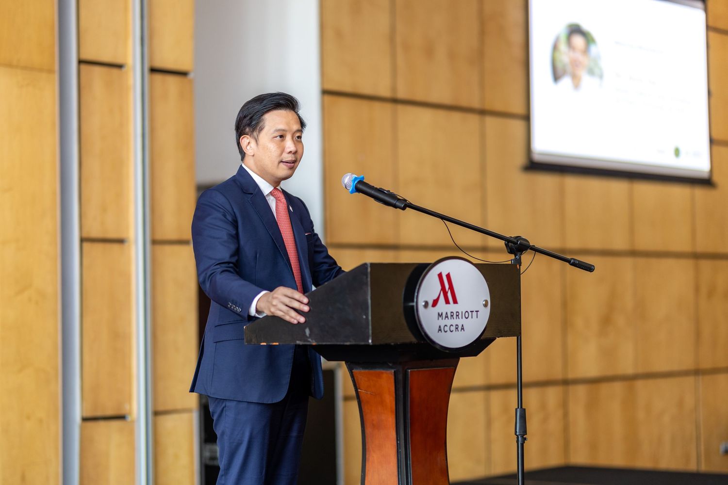 Man in navy suit speaking at Marriott Accra podium during professional conference event.