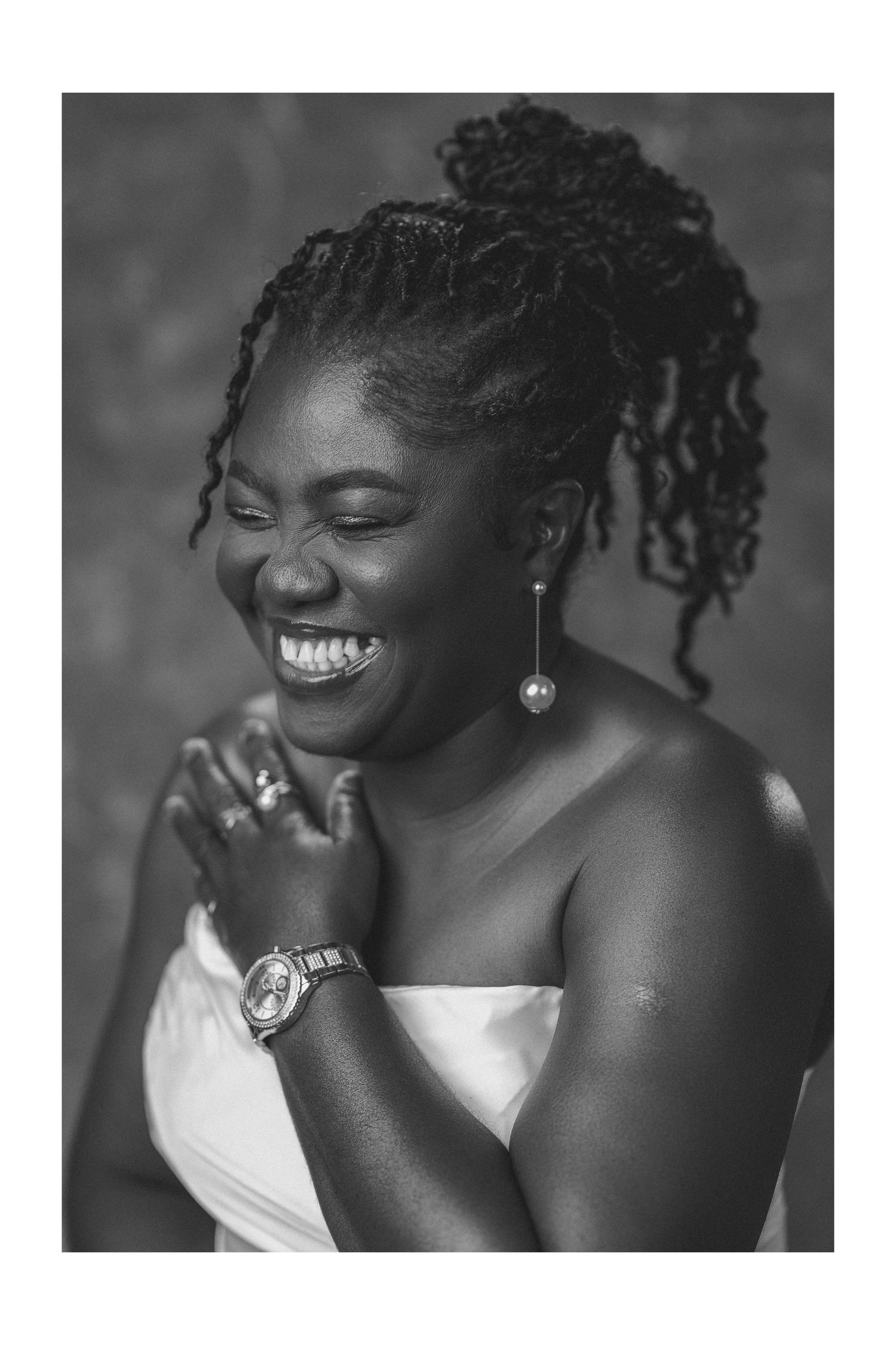 Black and white portrait of a laughing woman with braided updo, wearing a watch and earrings in a white dress.