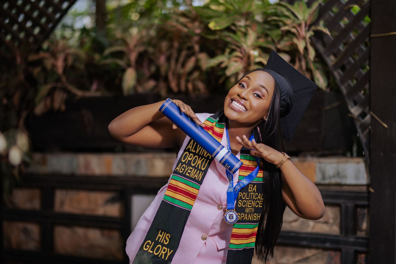 Joyful graduate in pink suit with kente stole and cap posing with diploma outdoors.