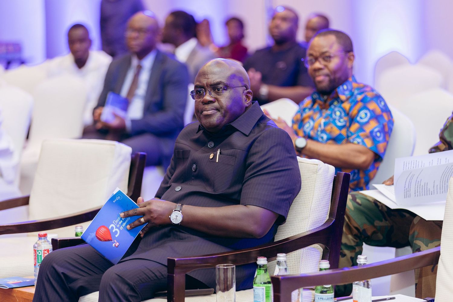 Smiling man in black attire seated at a conference holding a book with attendees in background.