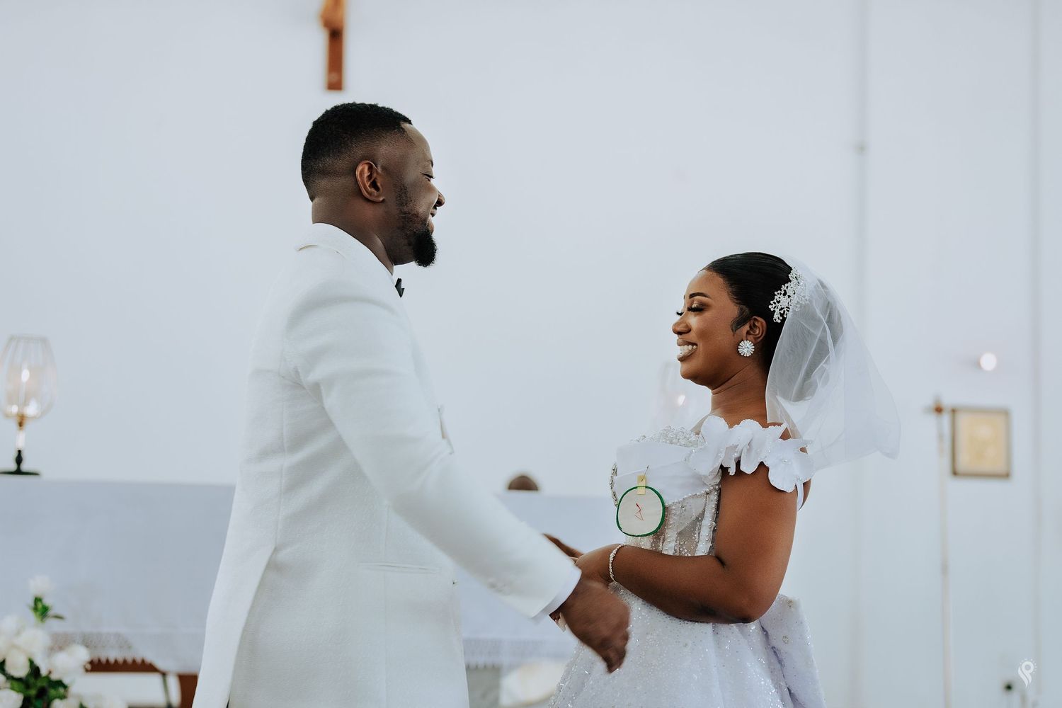 A joyful couple exchanges vows at a church wedding ceremony, with the bride in white lace and groom in a white suit.