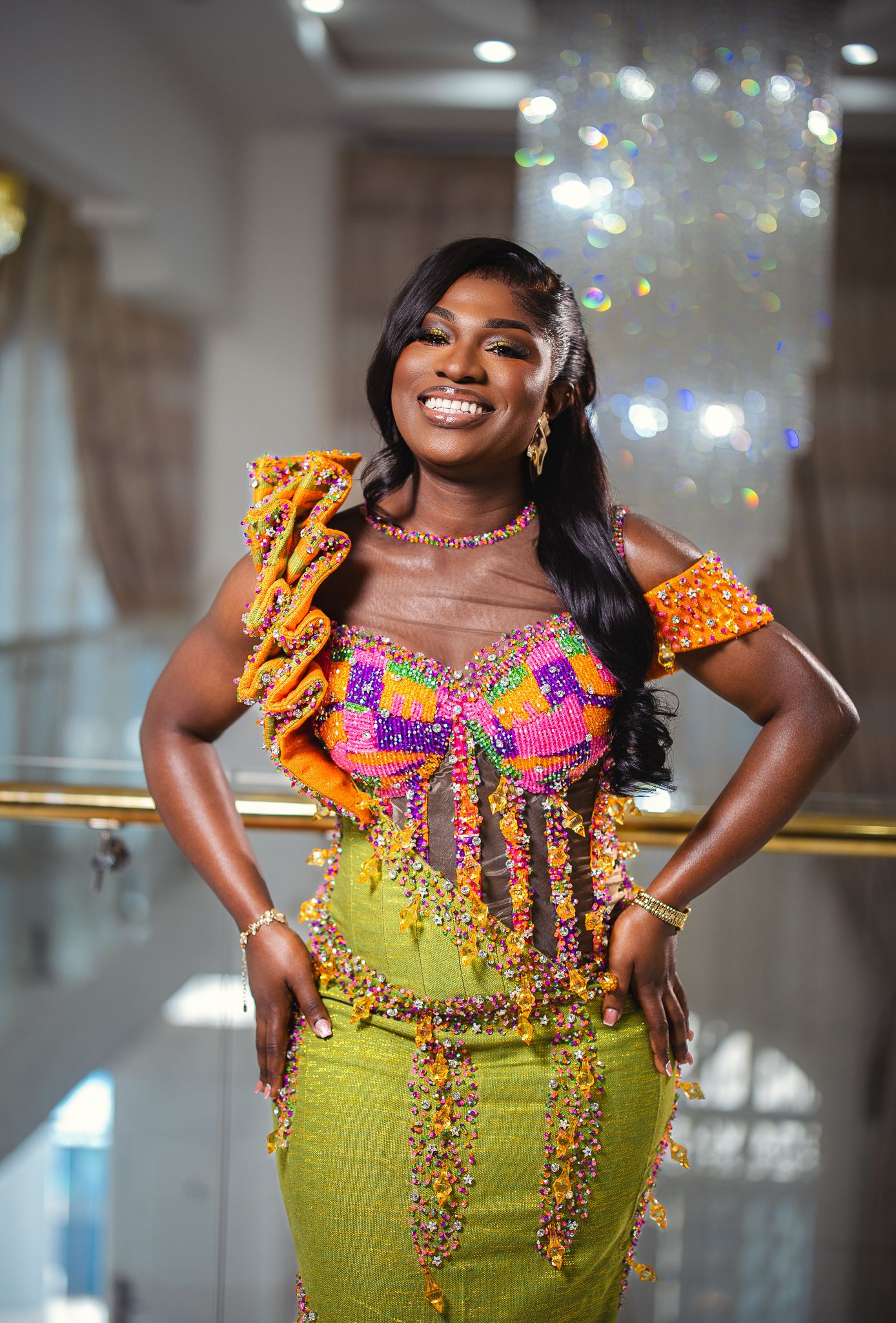 Woman in vibrant beaded Kente outfit smiling at a wedding celebration.