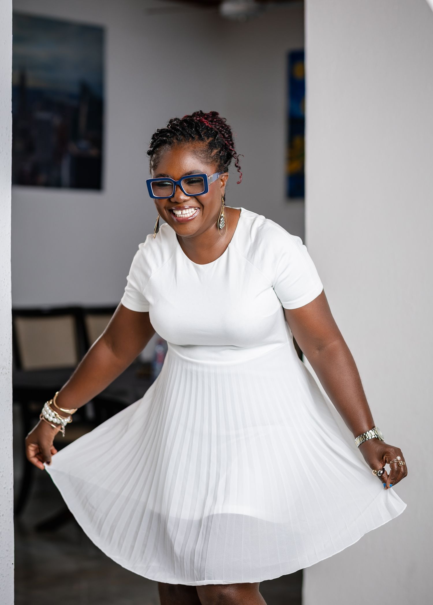 Smiling woman in white pleated dress and blue glasses twirling in a bright modern interior space.