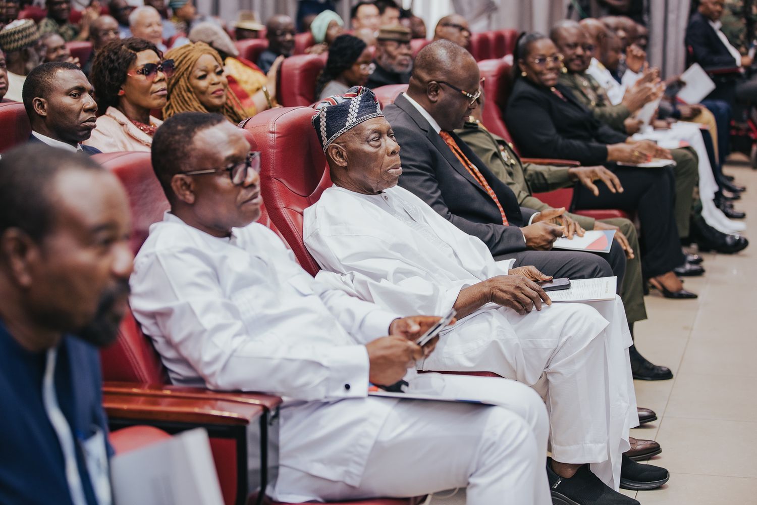 Attendees in white attire seated in red chairs at a formal conference event.