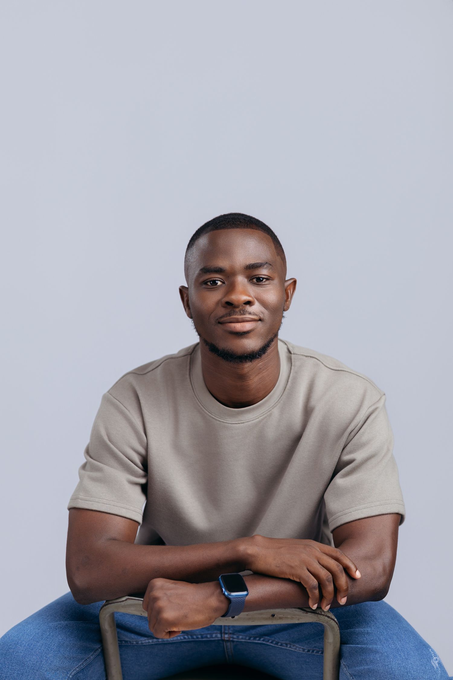 Young man in beige t-shirt and jeans sitting relaxed on chair against light gray studio background.