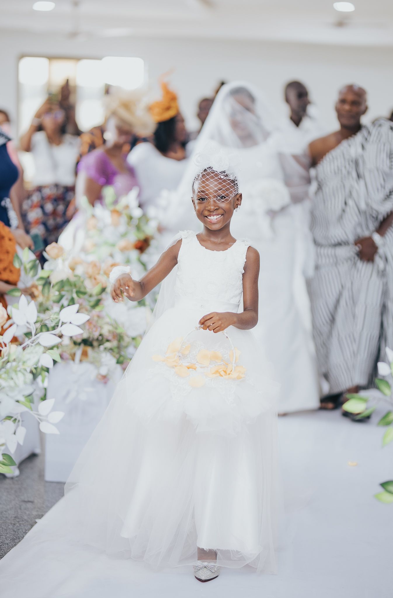 Adorable flower girl in white tulle dress scatters petals down the aisle at an elegant wedding ceremony.