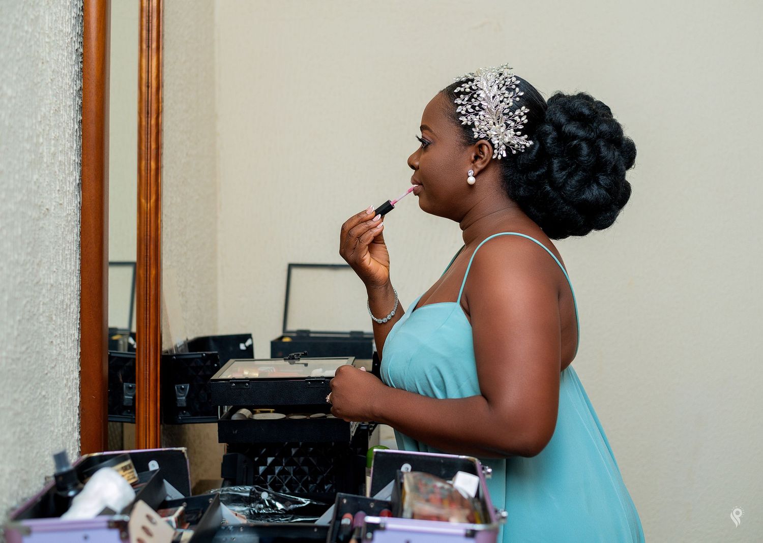 A bridesmaid in a teal dress applies lipstick while looking in a mirror during wedding preparations.