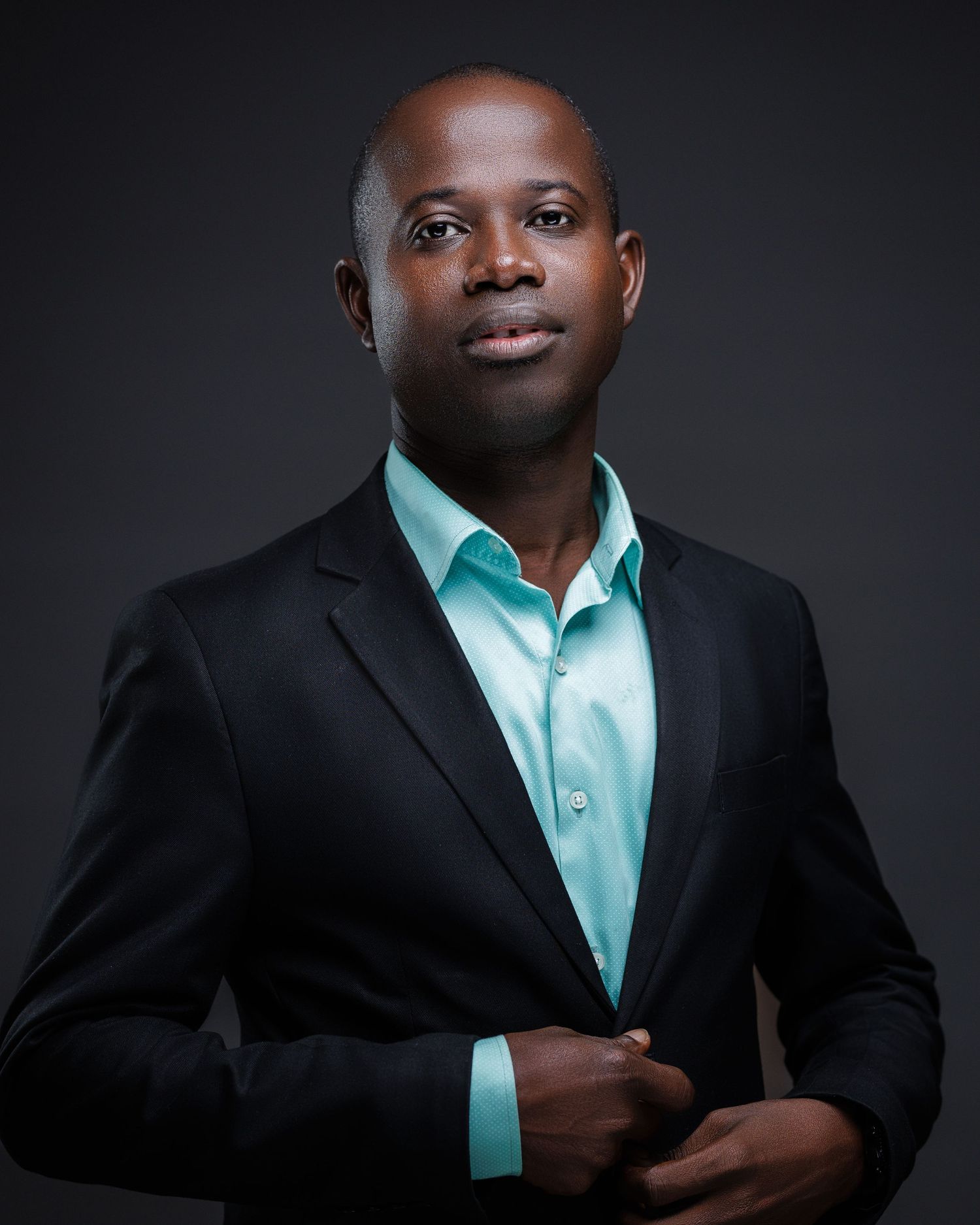 Young man in black blazer and teal shirt posing in sleek professional studio portrait.