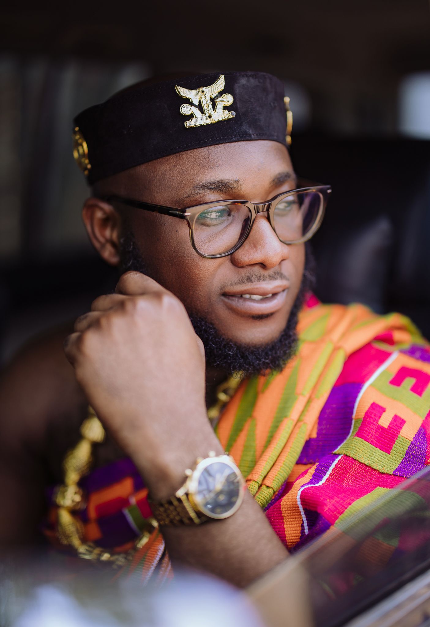 Man in traditional Kente cloth and black cap with gold accessories.