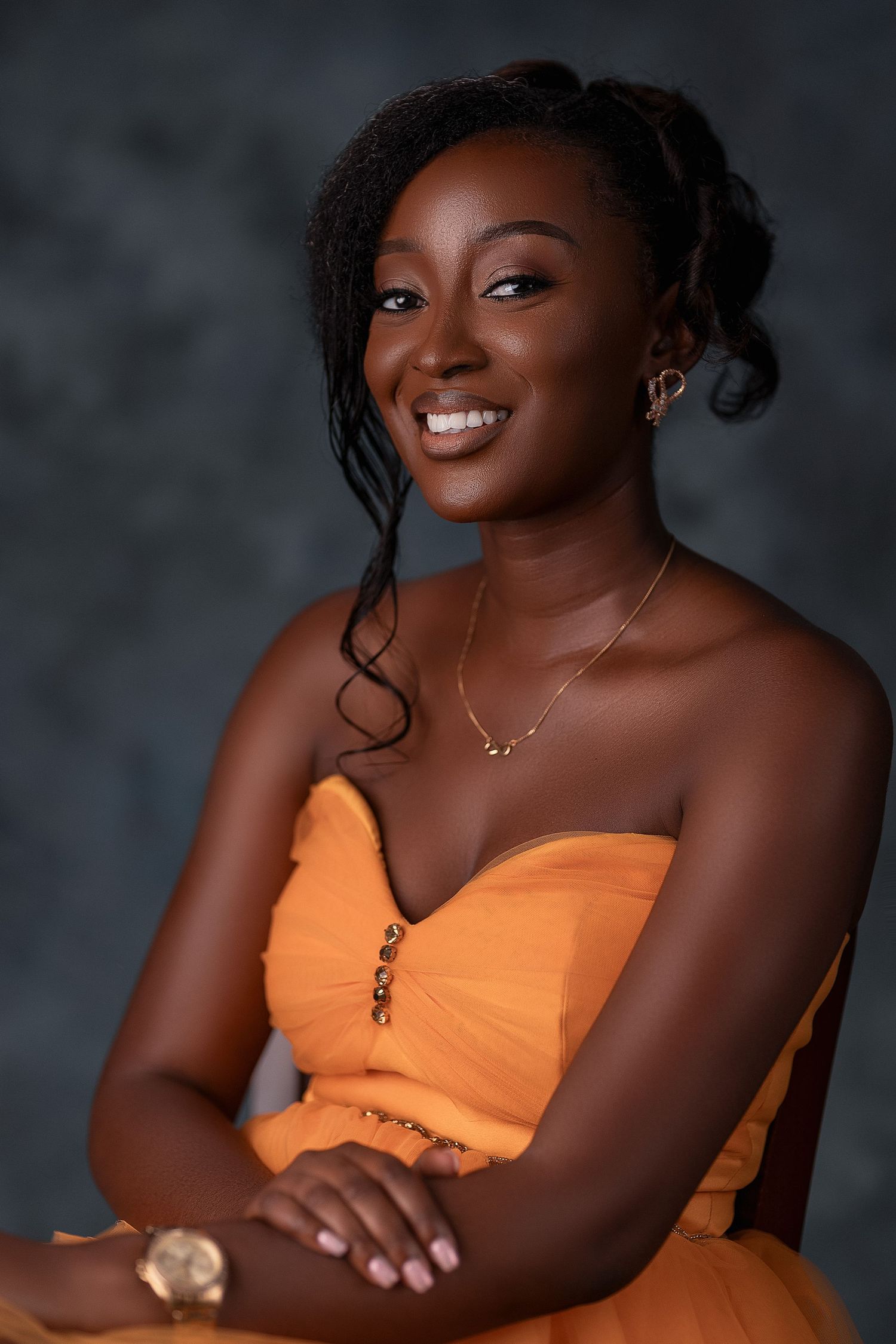 Smiling Black woman in orange strapless dress with gold jewelry seated elegantly.