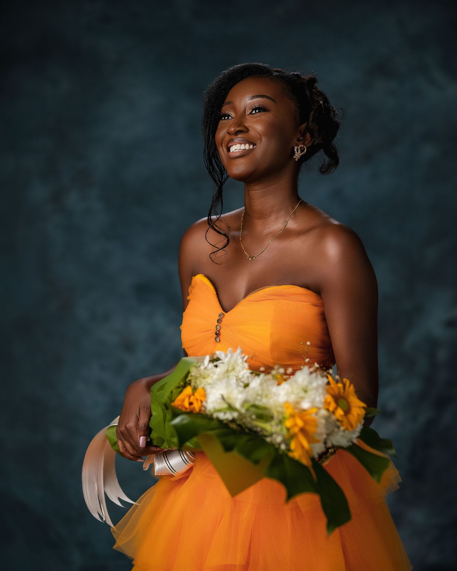 Young woman in strapless orange tulle gown holding sunflower bouquet, smiling against dark backdrop.