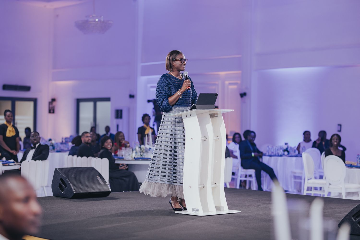 Woman in blue patterned dress speaks at podium during elegant evening banquet event.