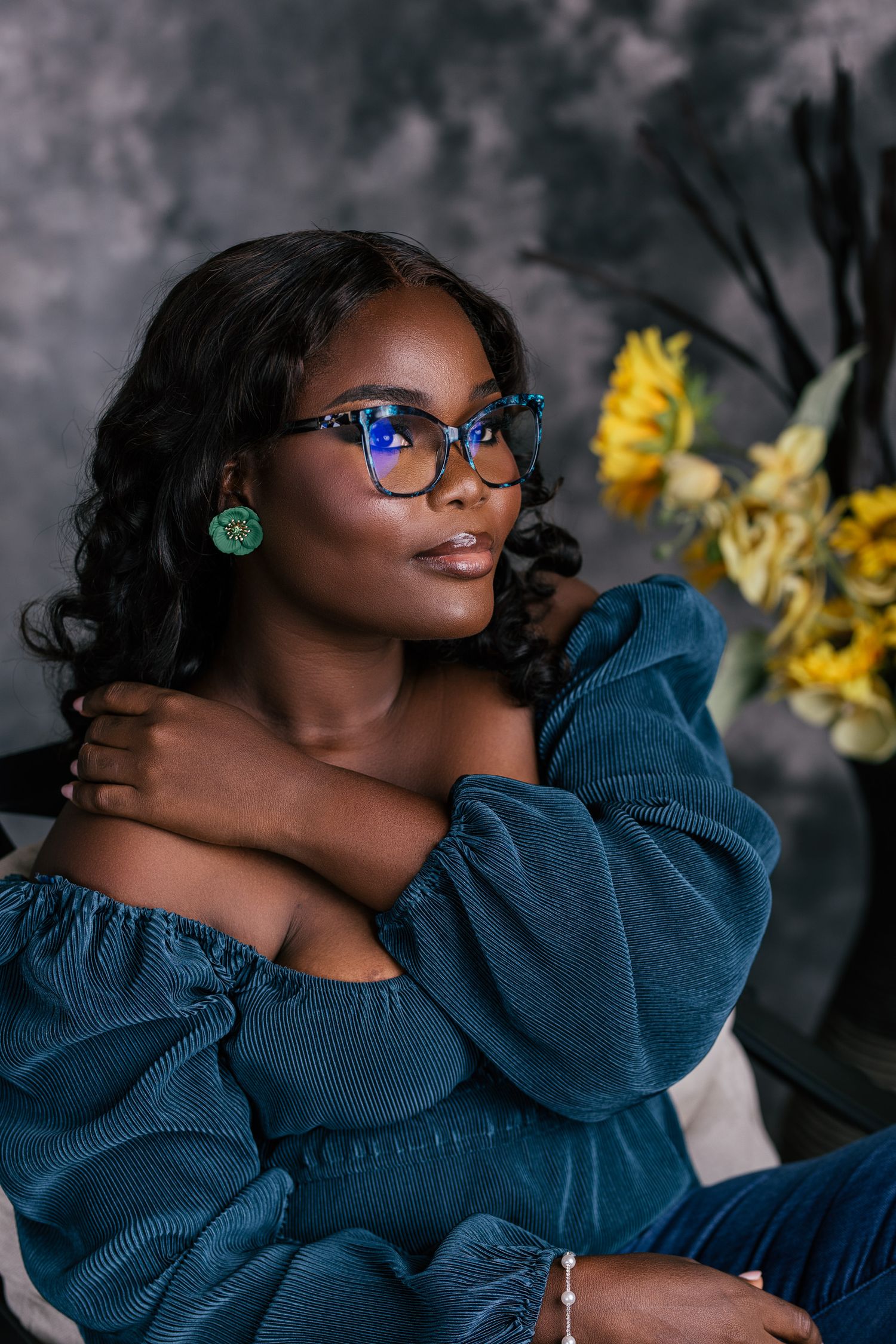 Beautiful woman in teal off-shoulder dress with green earrings and glasses posed near sunflowers in studio.