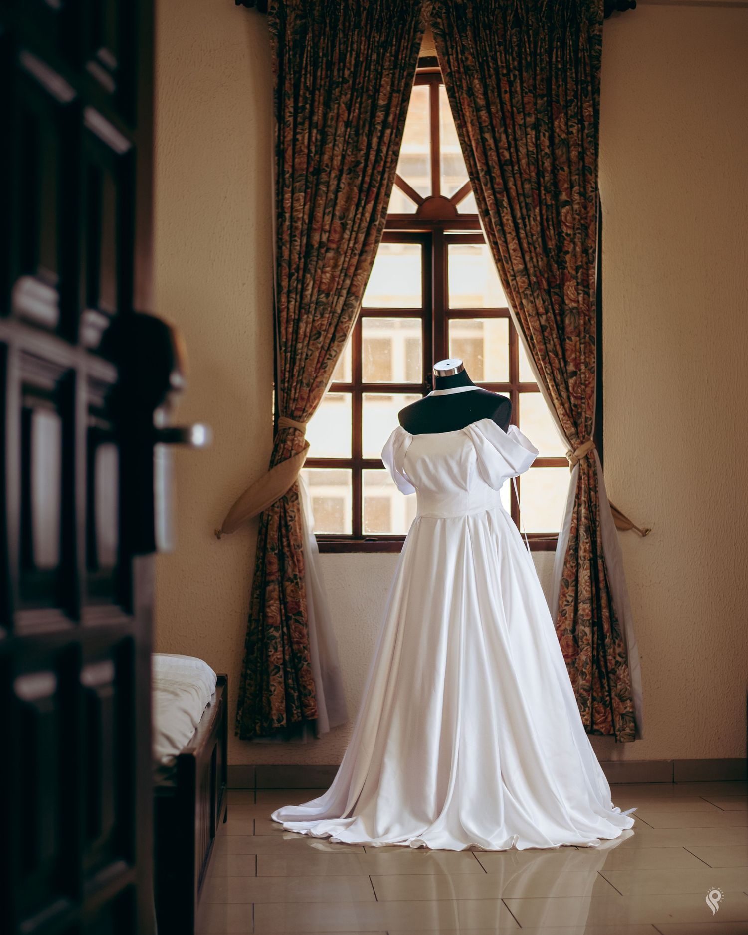 A white off-shoulder wedding gown displayed on a mannequin in front of a window with floral curtains.