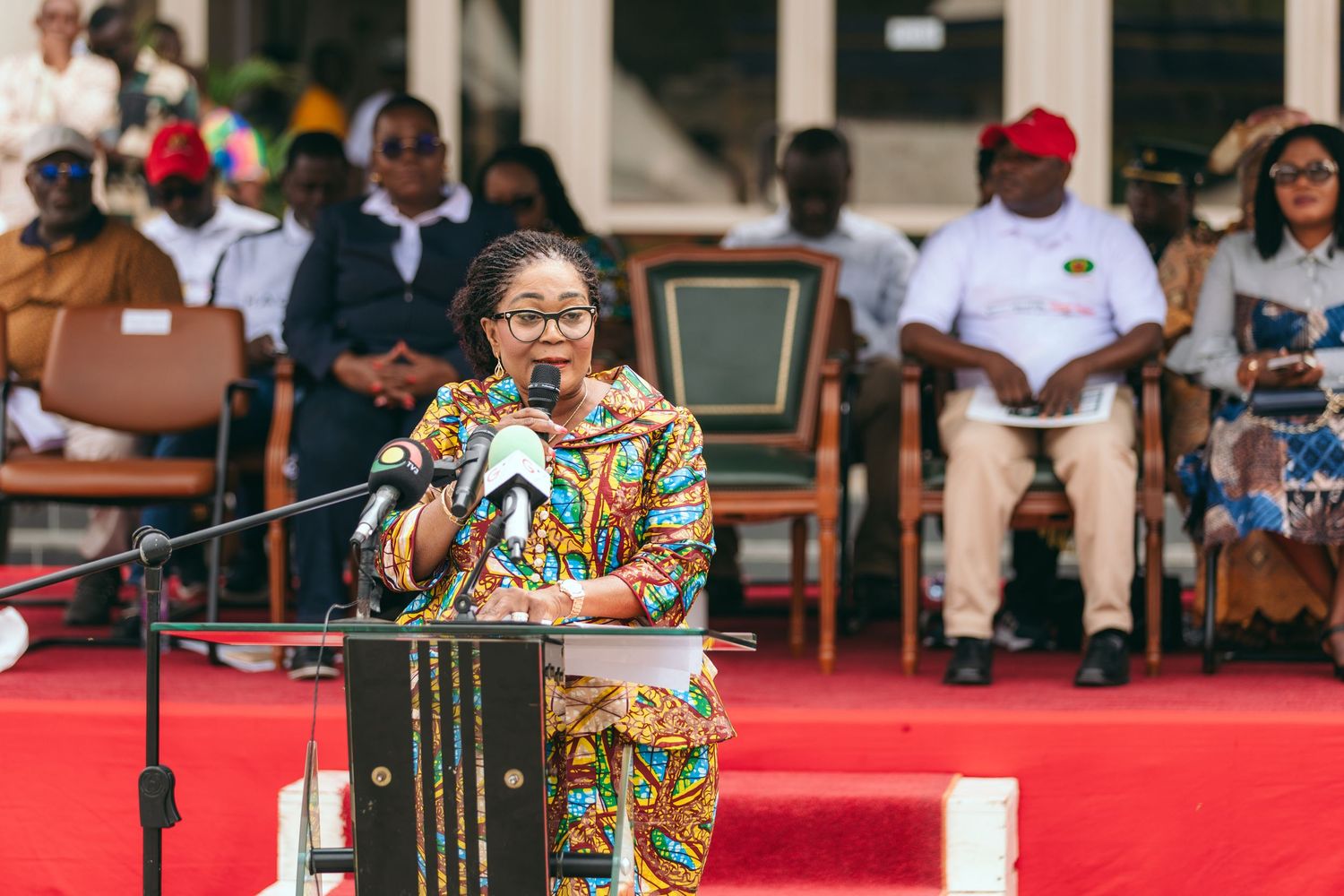 Woman in colorful kente cloth dress delivering speech at podium during outdoor cultural ceremony.