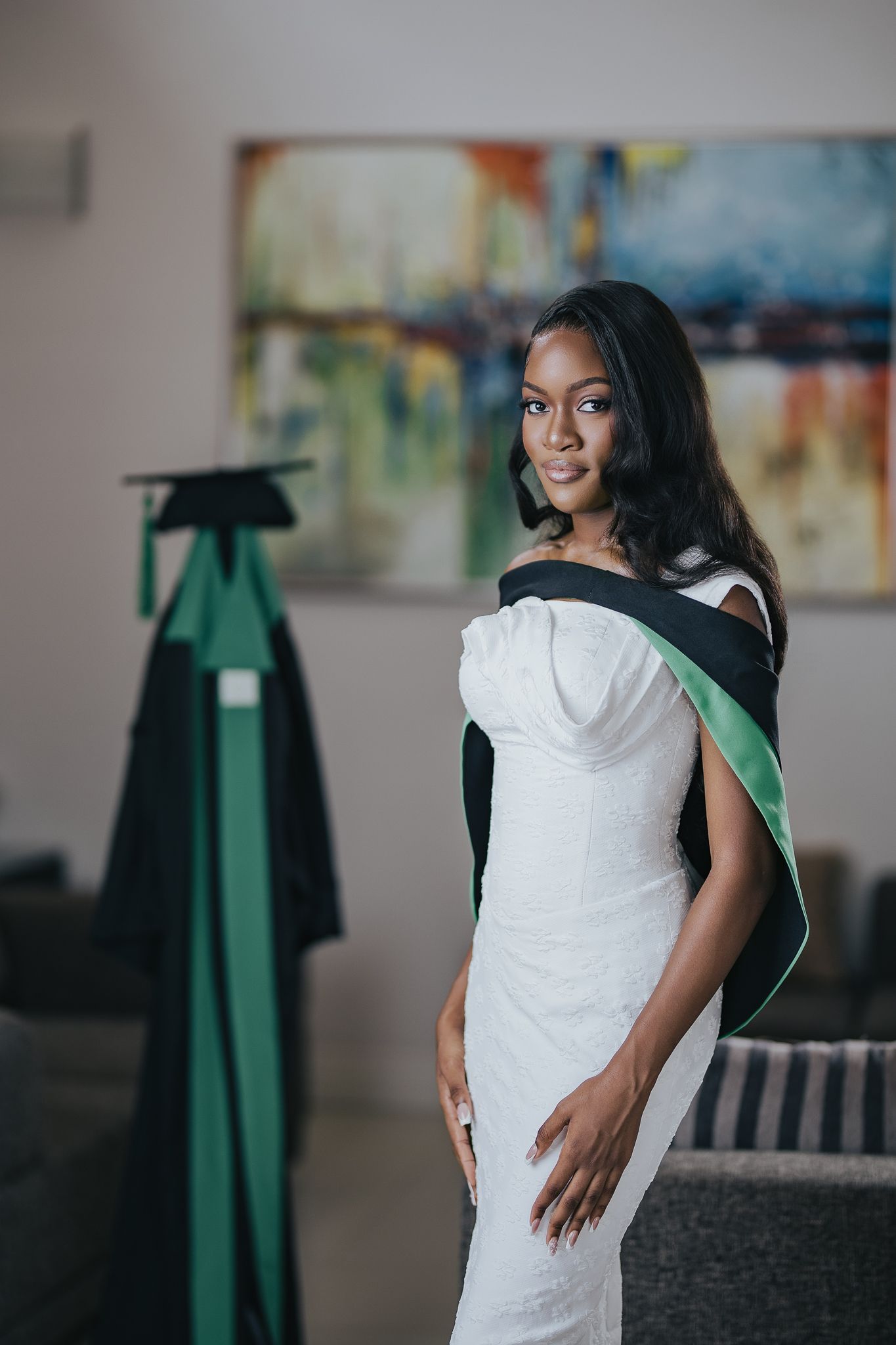Graceful woman in white fitted dress with green and black graduation sash posing indoors before ceremony.
