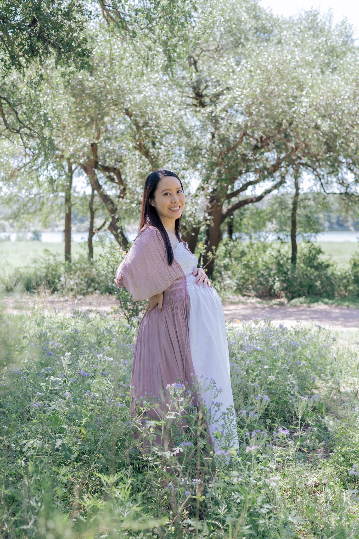 Pregnant mother expecting her third child is smiling at the camera in a park filled with lush green trees