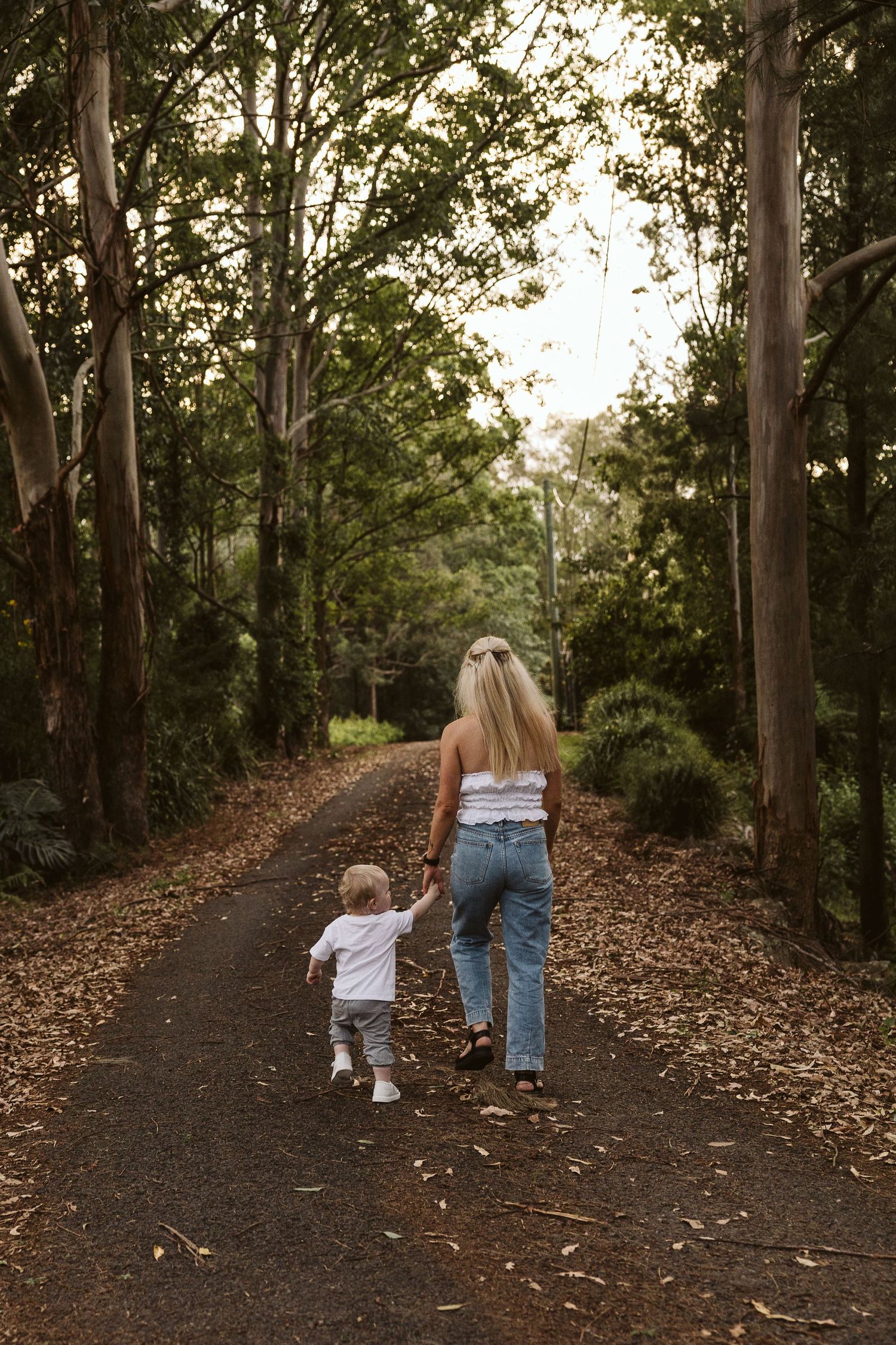 Mother and toddler walking hand in hand down a leafy tree-lined path in nature.