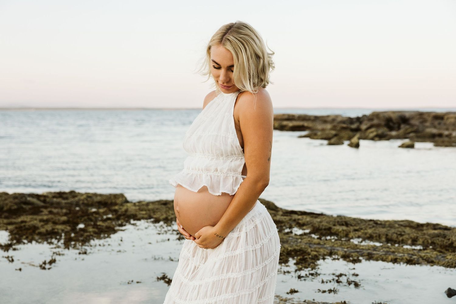 Pregnant blonde woman in white halter dress looking down at her baby bump on a serene rocky beach.