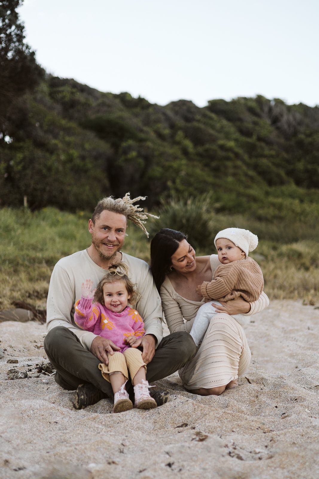 Happy family of four sitting on sandy beach, parents smiling with two young daughters in natural light.